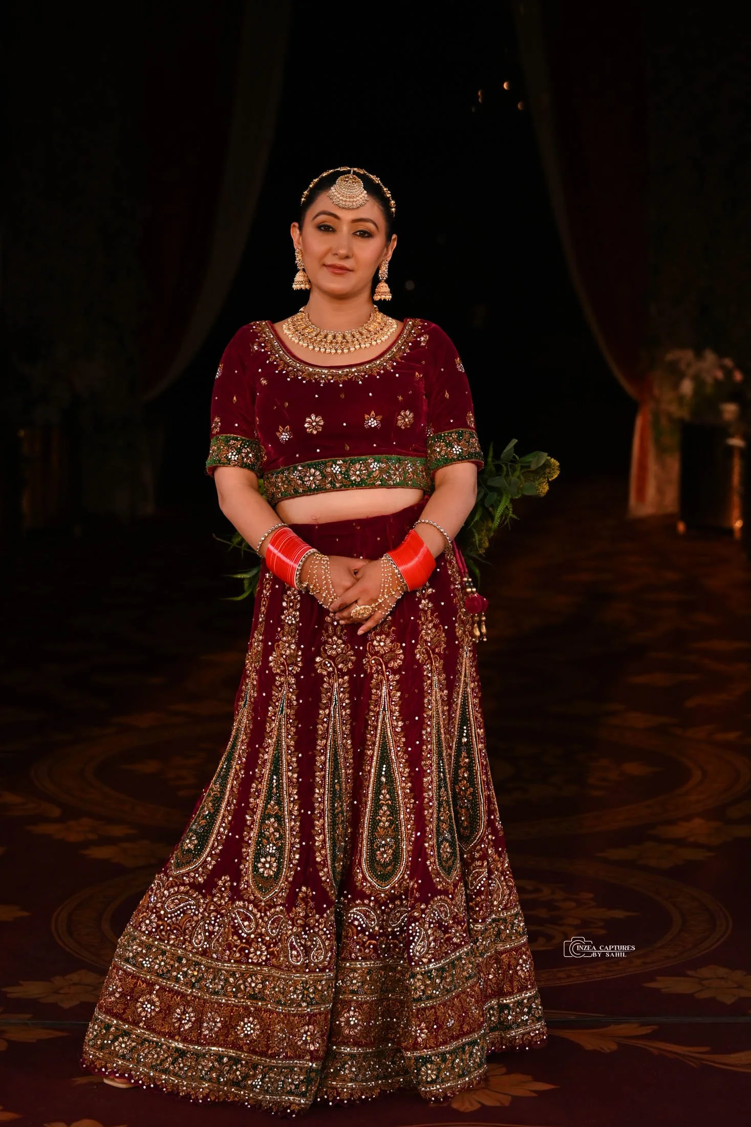 A woman wearing a traditional maroon Indian bridal lehenga with intricate embroidery and embellishments, paired with jewelry including earrings, necklace, and headpiece, standing on a decorated stage with a dark background.