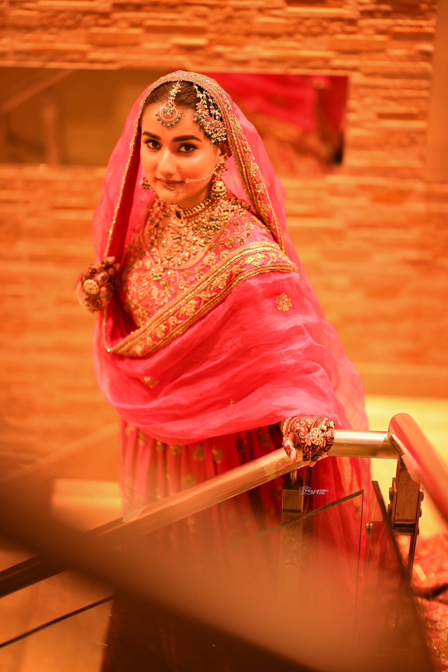 A woman dressed in traditional Indian attire, wearing a pink and gold embroidered saree, decorated with jewelry, including a nose ring, necklace, earrings, and headpiece, standing on a staircase with a warm brick wall background.