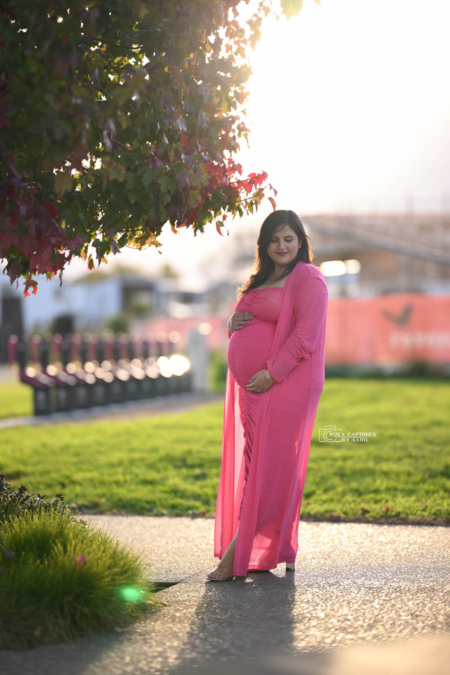 A pregnant woman in a pink dress and sheer cover-up standing on a pathway in a park, smiling and softly holding her belly, with a tree and sunset in the background.