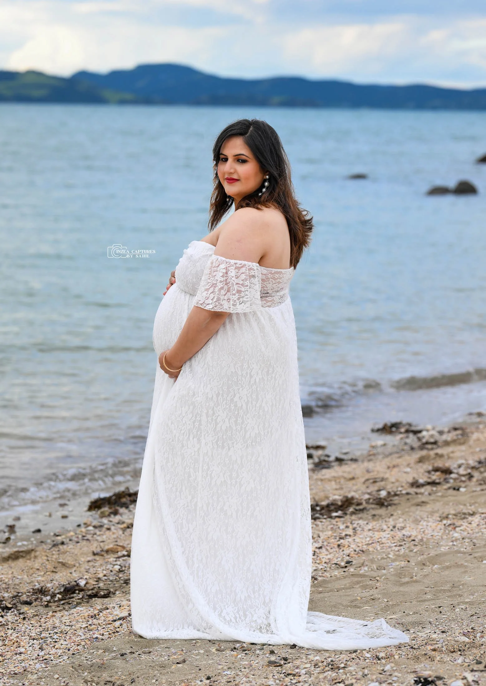 Pregnant woman in a white lace off-shoulder dress standing on a beach with water and mountains in the background.