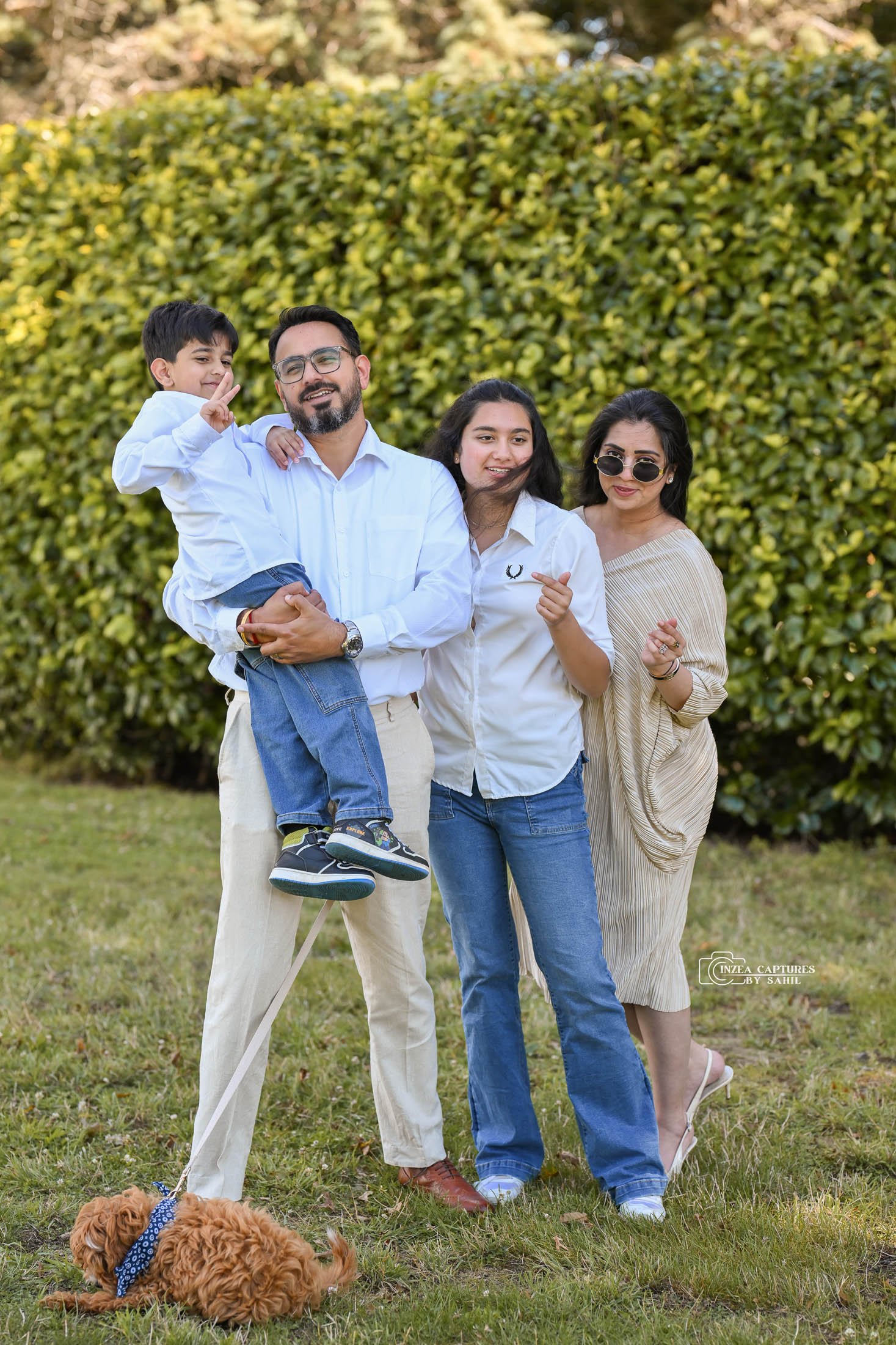 A family of four standing on grass outdoors in front of a large green bush, with a small brown dog lying on the ground nearby. The father holds a young boy in his arms, both smiling. Two women stand beside them, one with sunglasses and a striped dres