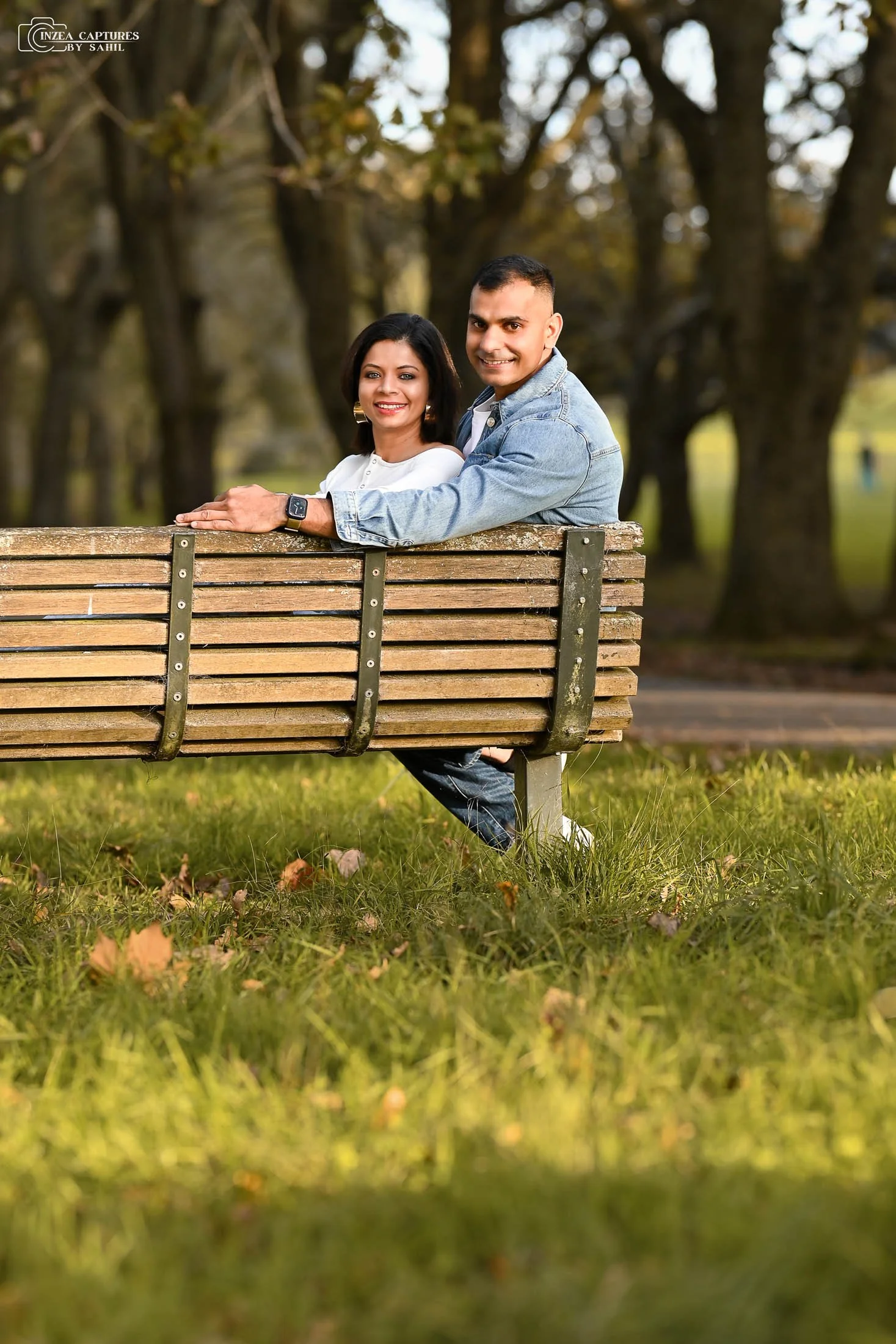 A couple sitting on a park bench outdoors, smiling at the camera with trees in the background.