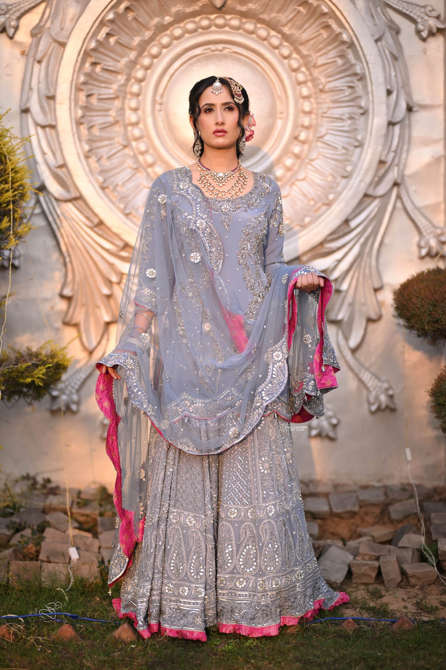 A woman dressed in a traditional silver and pink embroidered outfit, wearing jewelry, standing outdoors in front of an elaborate decorative wall with ornate patterns and surrounded by greenery.