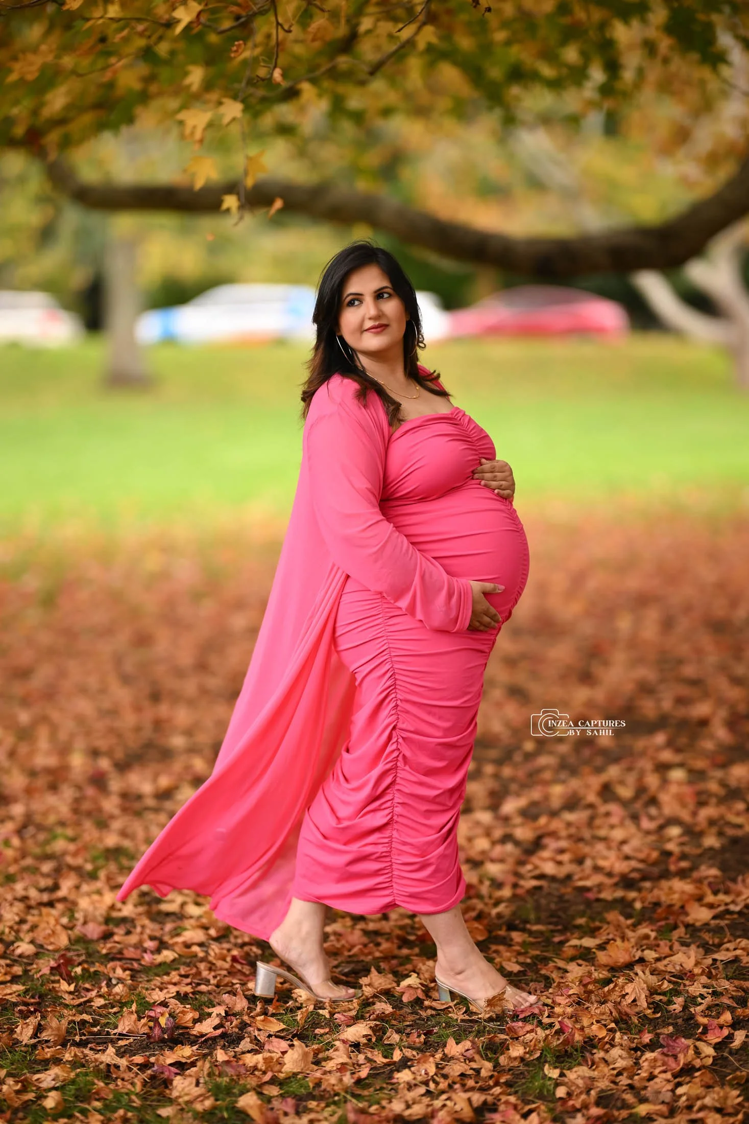 A pregnant woman in a pink dress and matching shawl standing outdoors on fallen autumn leaves, with trees and parked cars in the background.