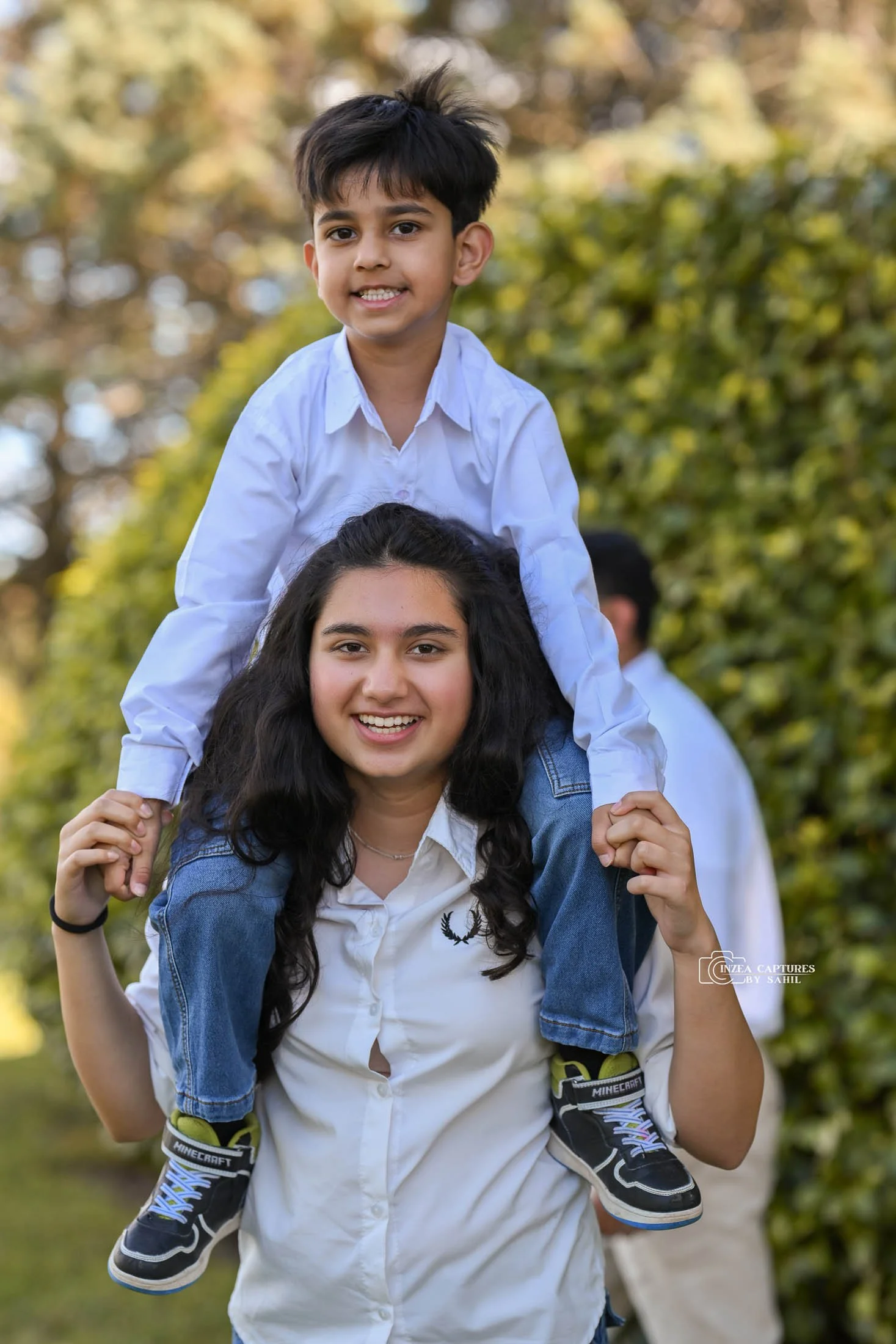 A girl with long dark hair and a white shirt is smiling while giving a piggyback ride to a boy with short dark hair, wearing a white shirt and blue jeans, outdoors in a park with green and yellow foliage.