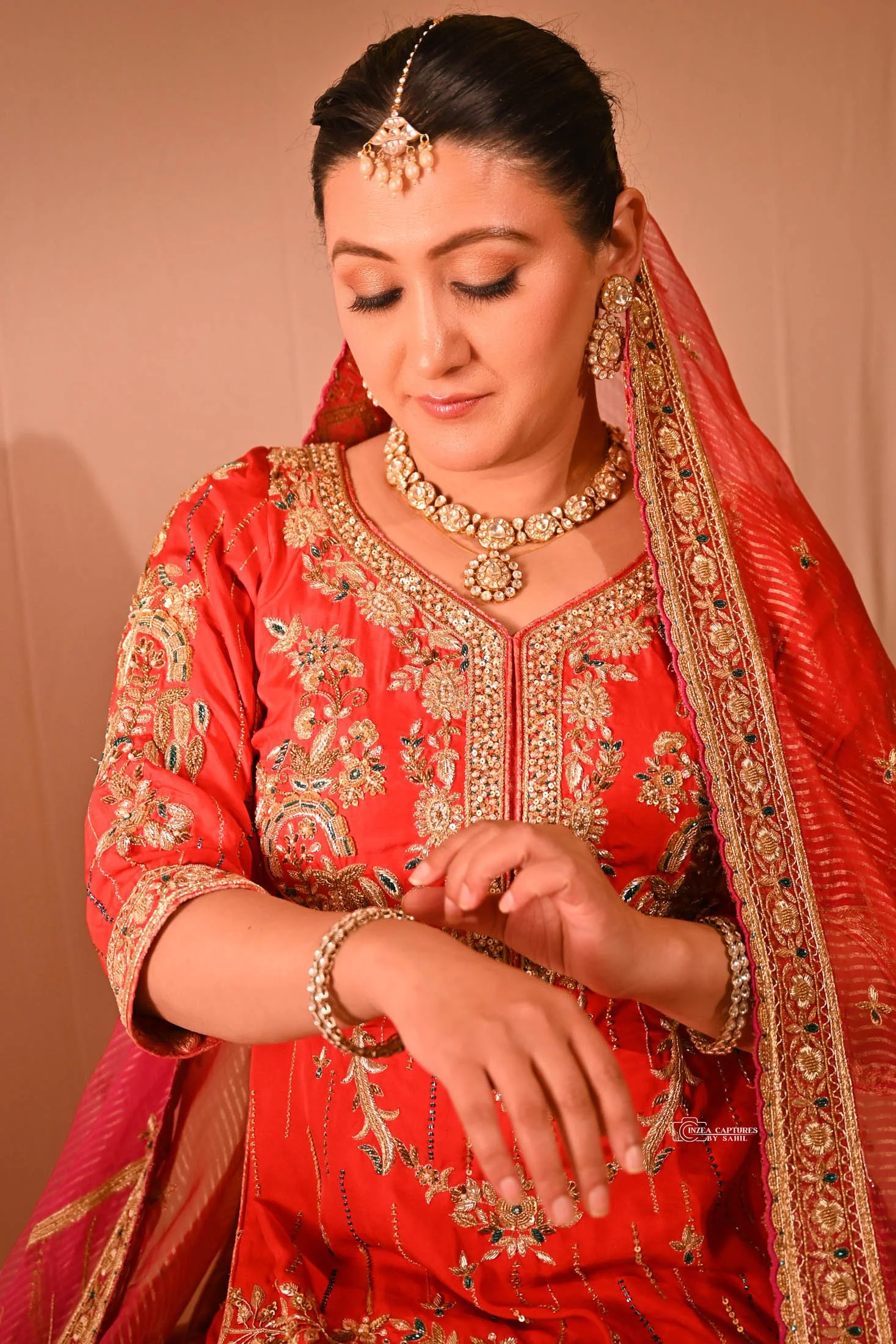 A woman in traditional Indian attire, wearing a red embroidered dress and gold jewelry, is putting on a bracelet.