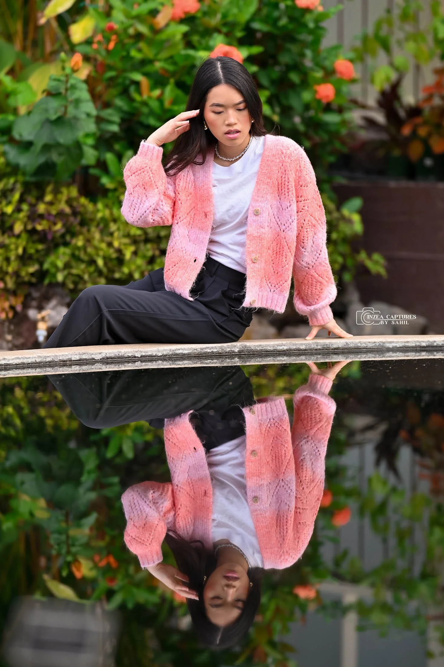 A woman in a pink knitted sweater, white shirt, and black pants sitting on the edge of a fountain, looking down with her hand near her ear, with her reflection visible in the water below, surrounded by green and orange plants.