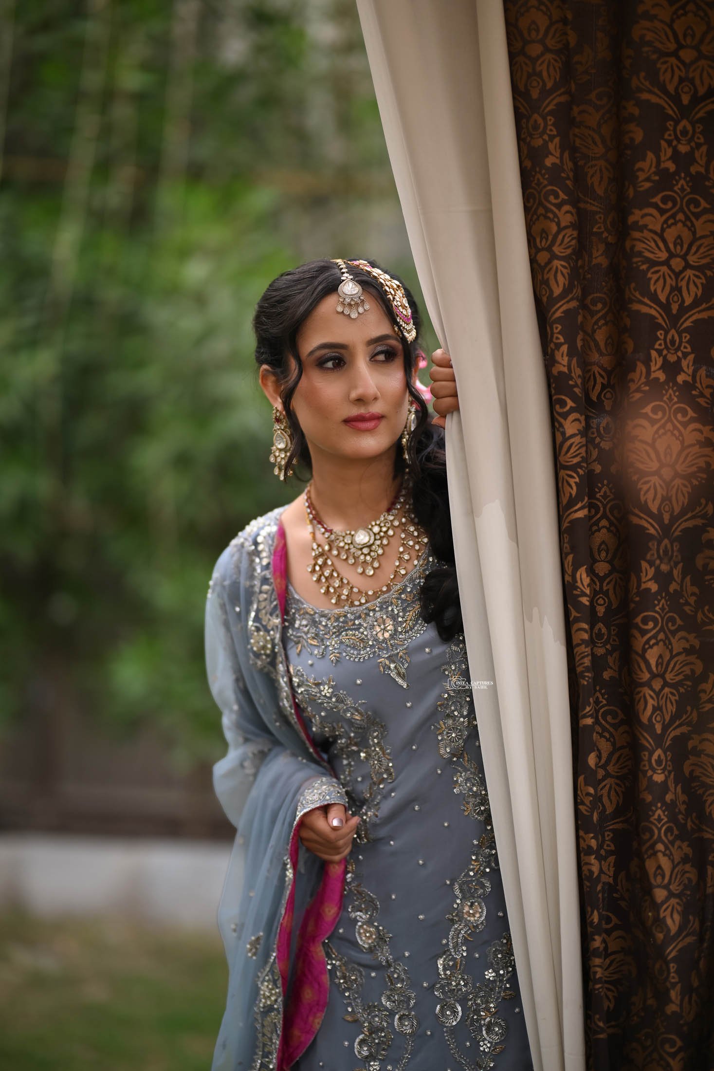 A woman dressed in traditional Indian attire peeking from behind a curtain, outdoors with green trees in the background.