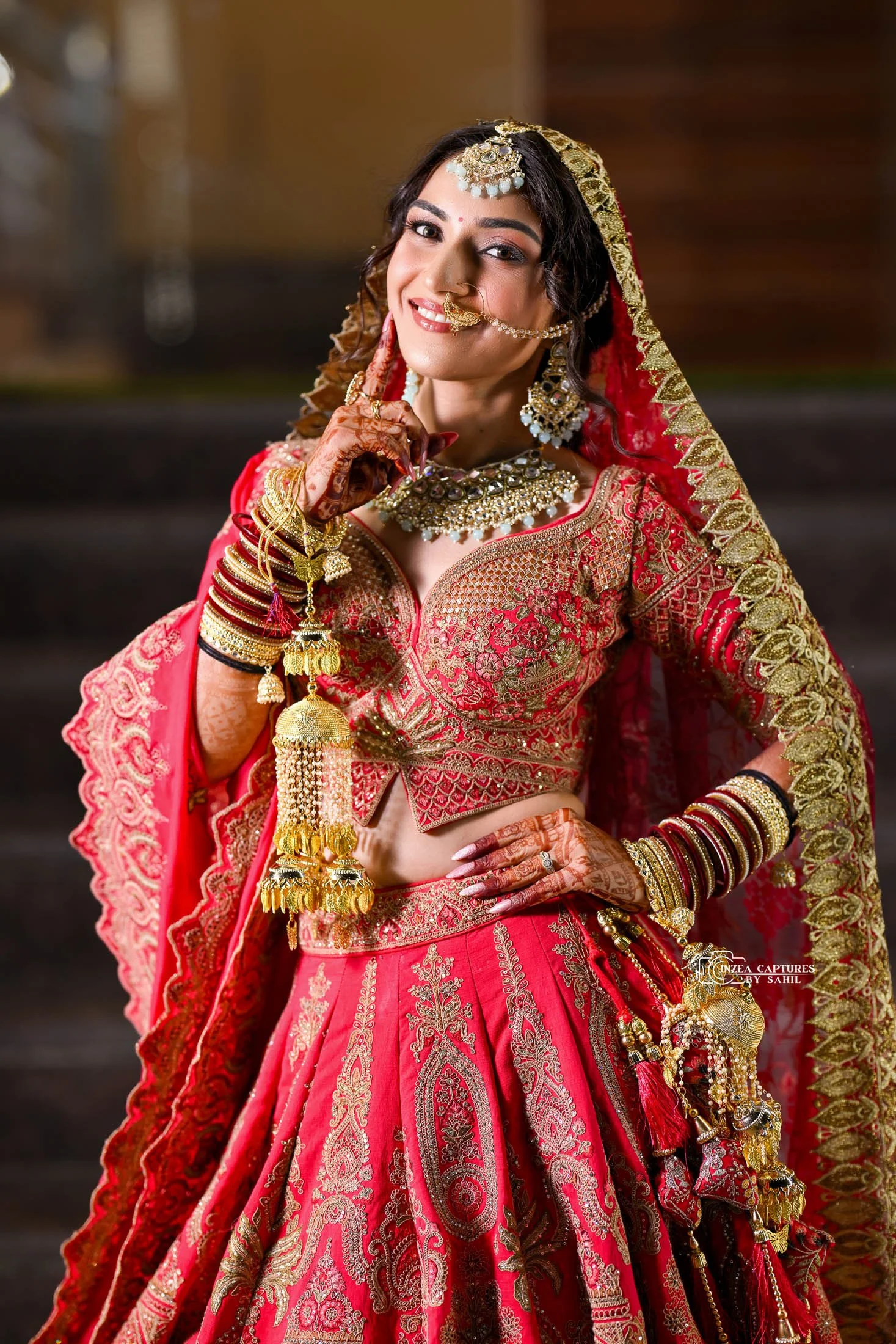 A woman dressed in traditional bridal Indian attire, wearing a red and gold embroidered lehenga, jewelry including earrings, necklace, bangles, nose ring, and headpiece, with henna on her hands, smiling and posing with one hand on her waist and the o