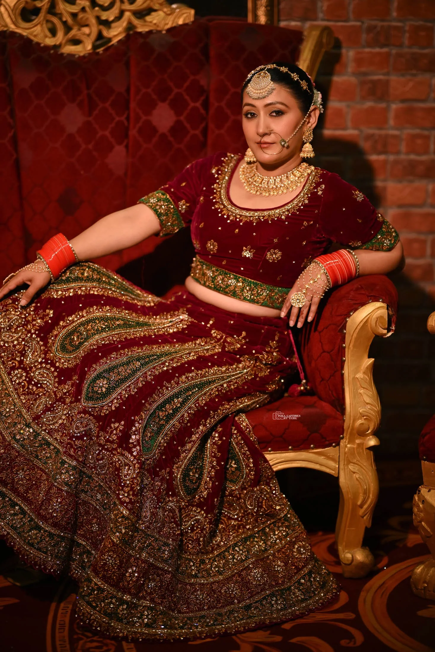 A woman wearing a traditional Indian red and gold heavily embroidered sari, gold jewelry, and a headpiece, sitting on a vintage red and gold velvet chair with a brick wall background.