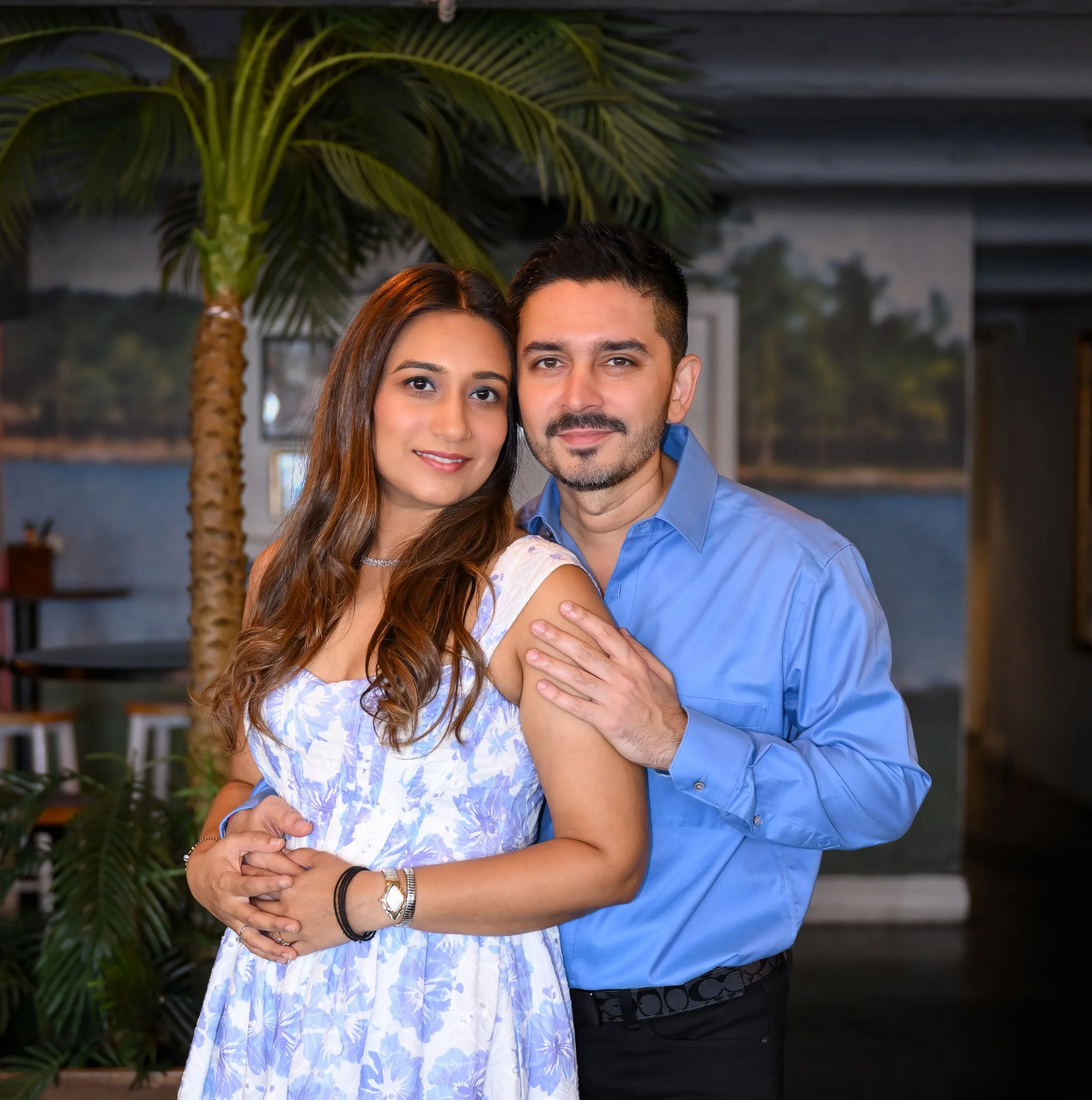 A couple posing together indoors, with a large palm plant and a lakeside mural in the background.