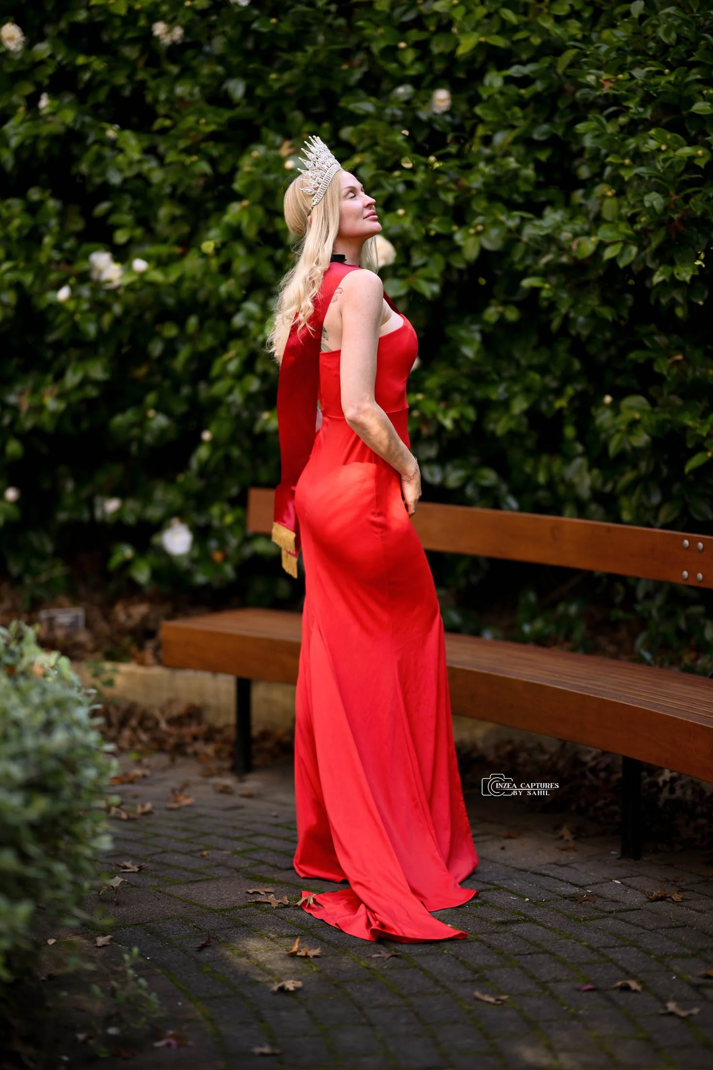 A woman wearing a red gown and a silver crown stands outdoors on a paved walkway, with green foliage in the background and a wooden bench nearby.