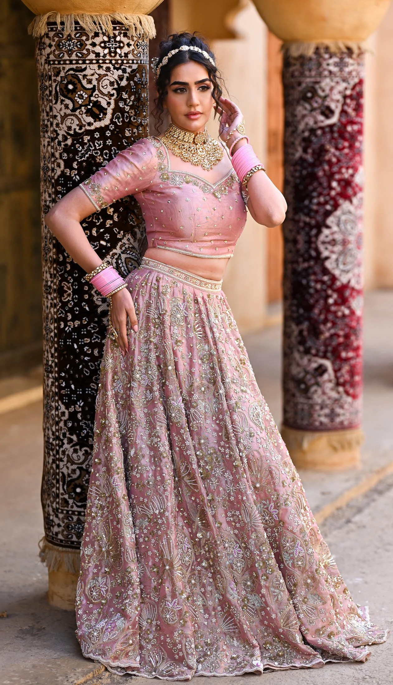 A woman in traditional Indian attire, wearing a pink embroidered lehenga, gold jewelry, and pink bangles, posing outdoors between decorated pillars.