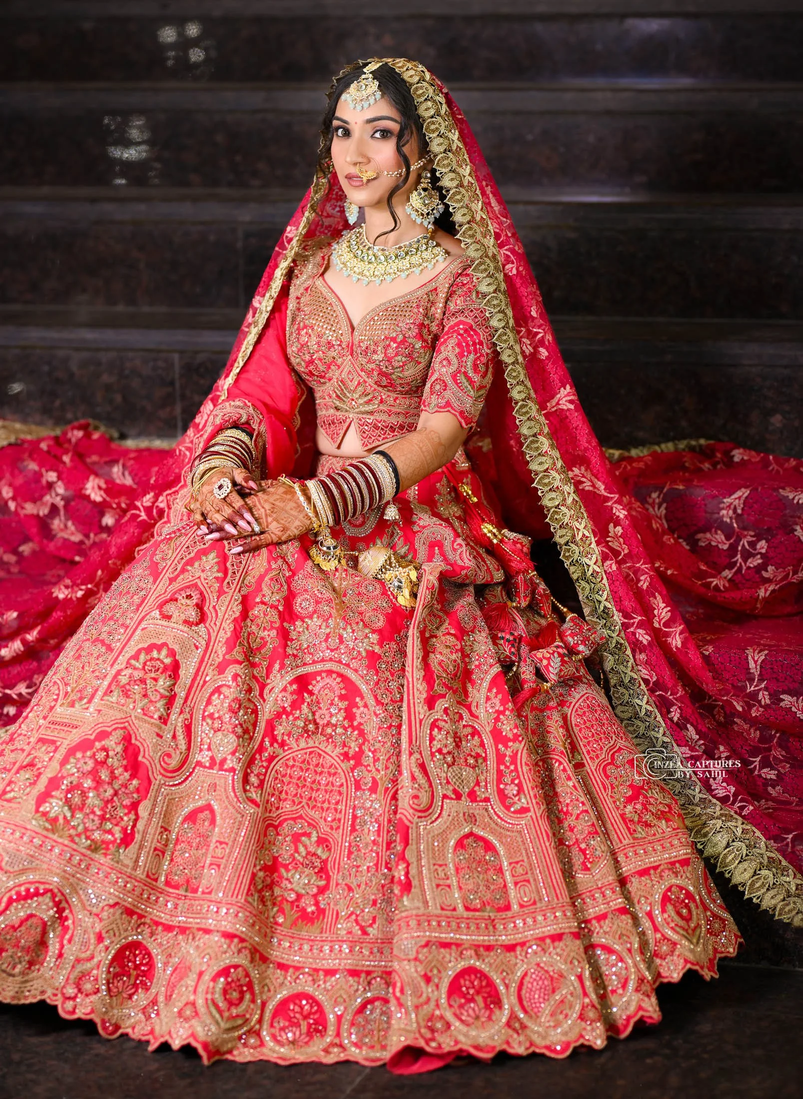 Indian bride wearing red and gold embroidered traditional bridal outfit with jewelry, including necklace, earrings, nose ring, bracelets, and headpiece, sitting on dark stairs.