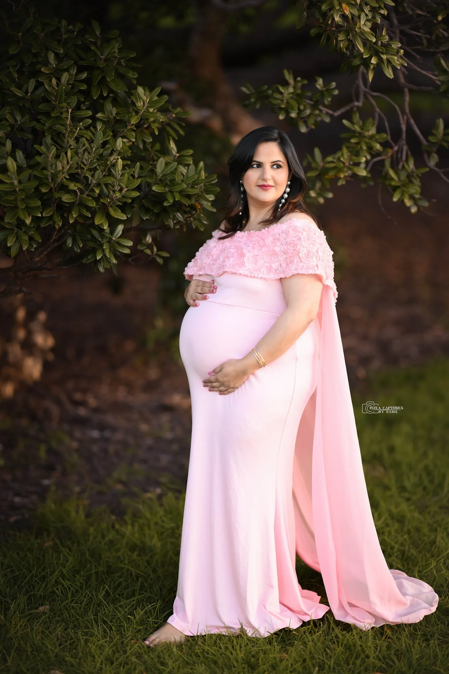 A pregnant woman in a pink dress standing outdoors near green foliage. She is holding her belly with both hands and looking to the side with a slight smile.