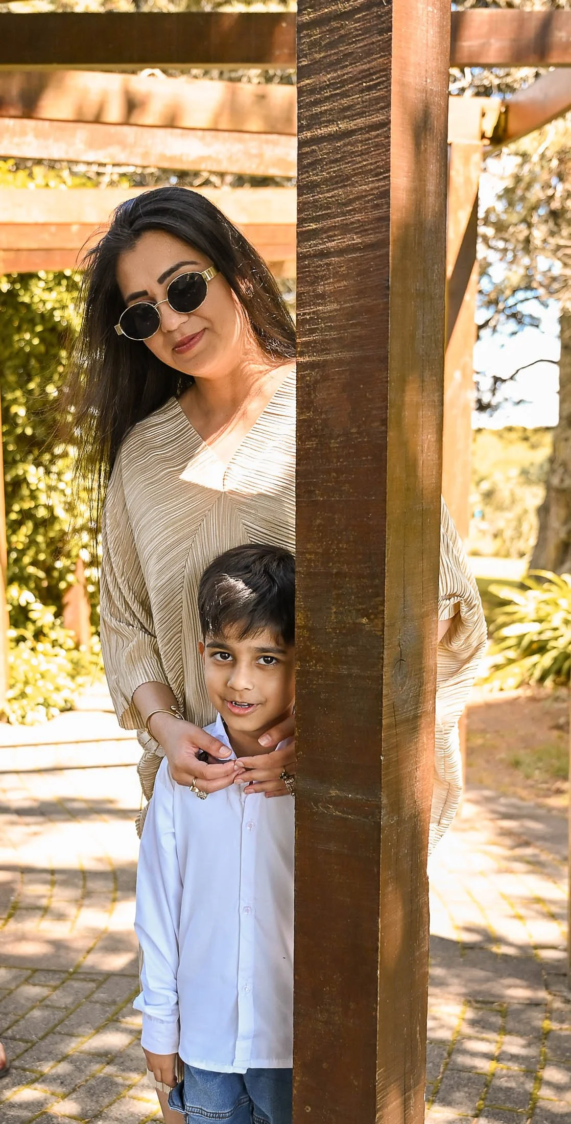 A woman with dark hair, wearing sunglasses and a beige blouse, stands behind a wooden post with a young boy in a white shirt and jeans, both peeking out from behind the post outdoors with greenery and trees in the background.