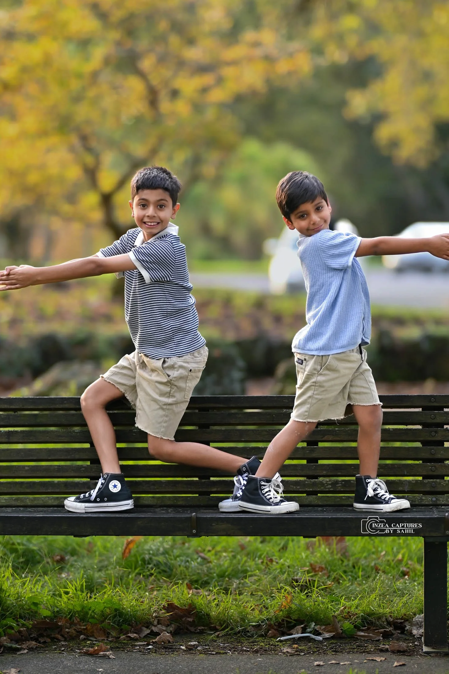 Two young boys with dark hair, wearing shorts, T-shirts, and sneakers, balancing and holding onto each other's outstretched arms while standing on a park bench with green grass and trees with yellow leaves in the background during fall.