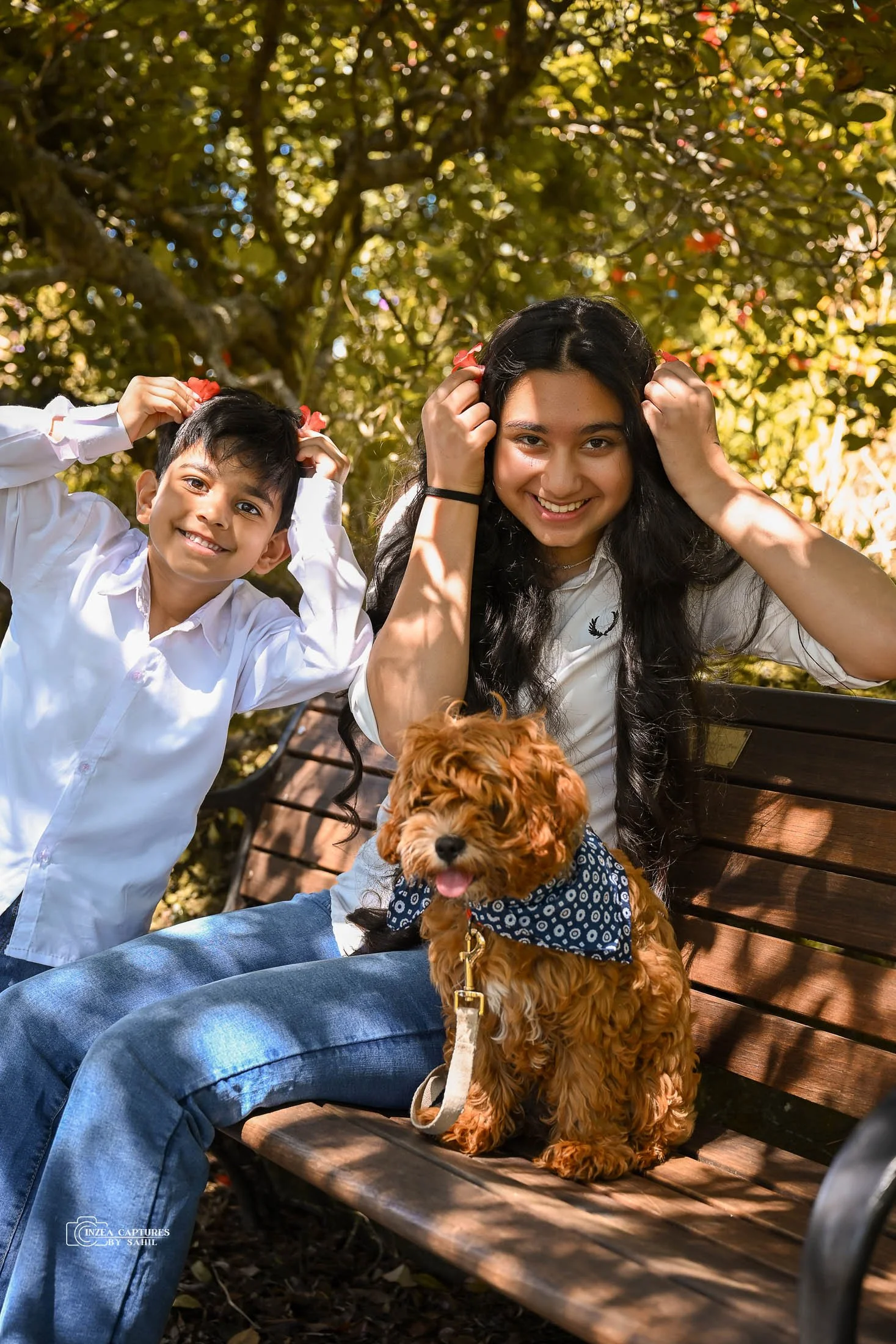 A woman and a young boy sit on a park bench with a fluffy brown dog wearing a blue bandana. They are outdoors under sunlight filtering through leaves, smiling and playing.