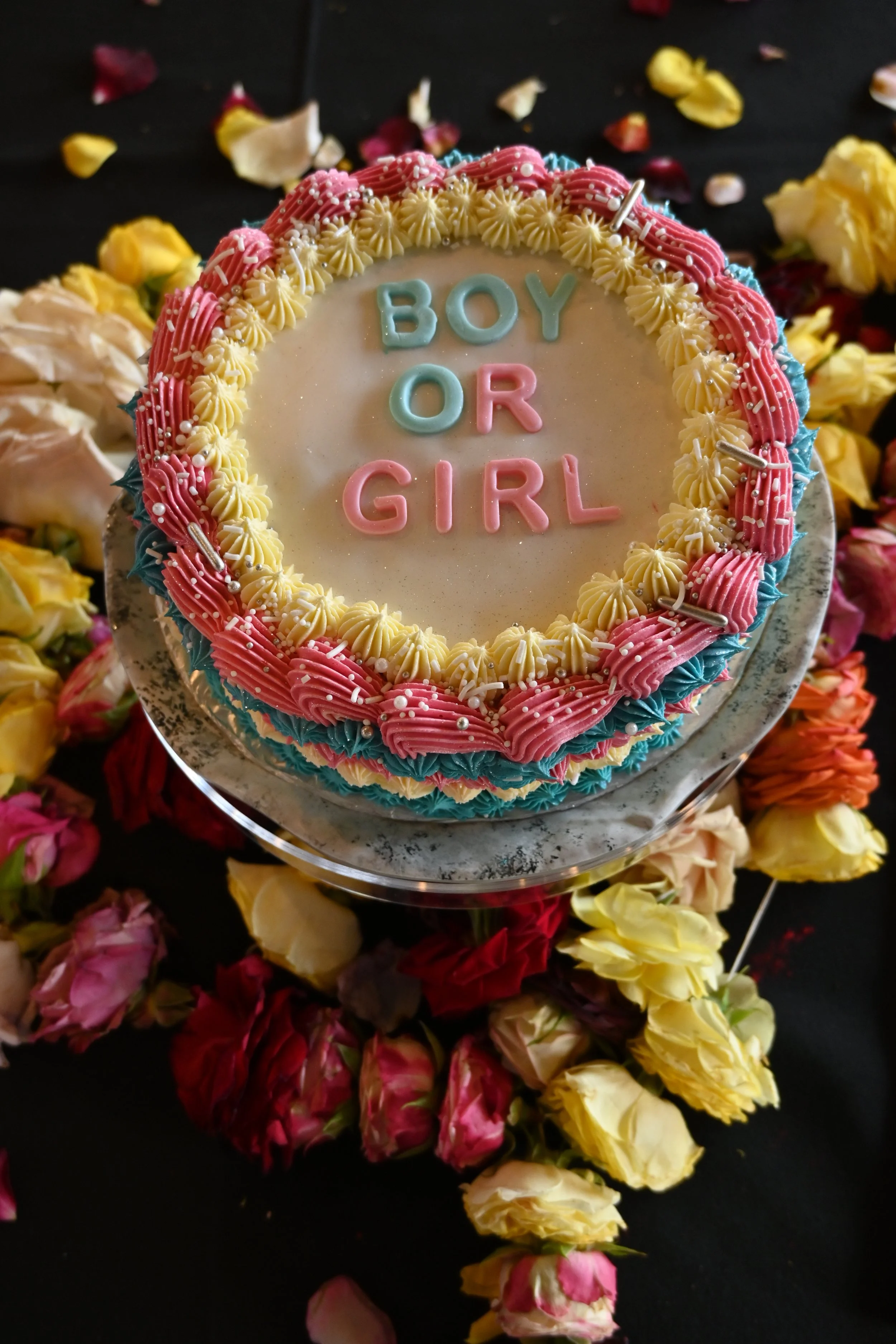 Decorated cake with the words 'Boy or Girl' written on it, surrounded by pink, yellow, and blue icing, placed on a cake stand amid scattered flowers.