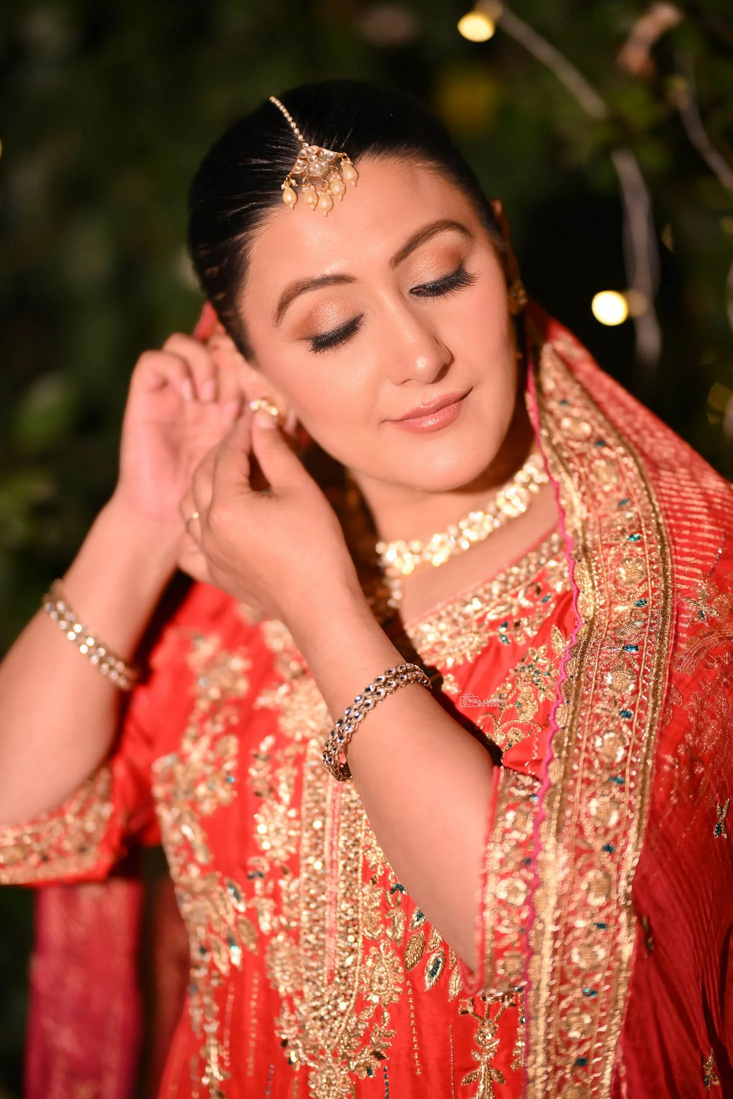 A woman wearing a red traditional Indian outfit with gold embroidery, adjusting her earrings with a serene expression, accessorized with gold jewelry and a pearl hair ornament, outdoors at night.