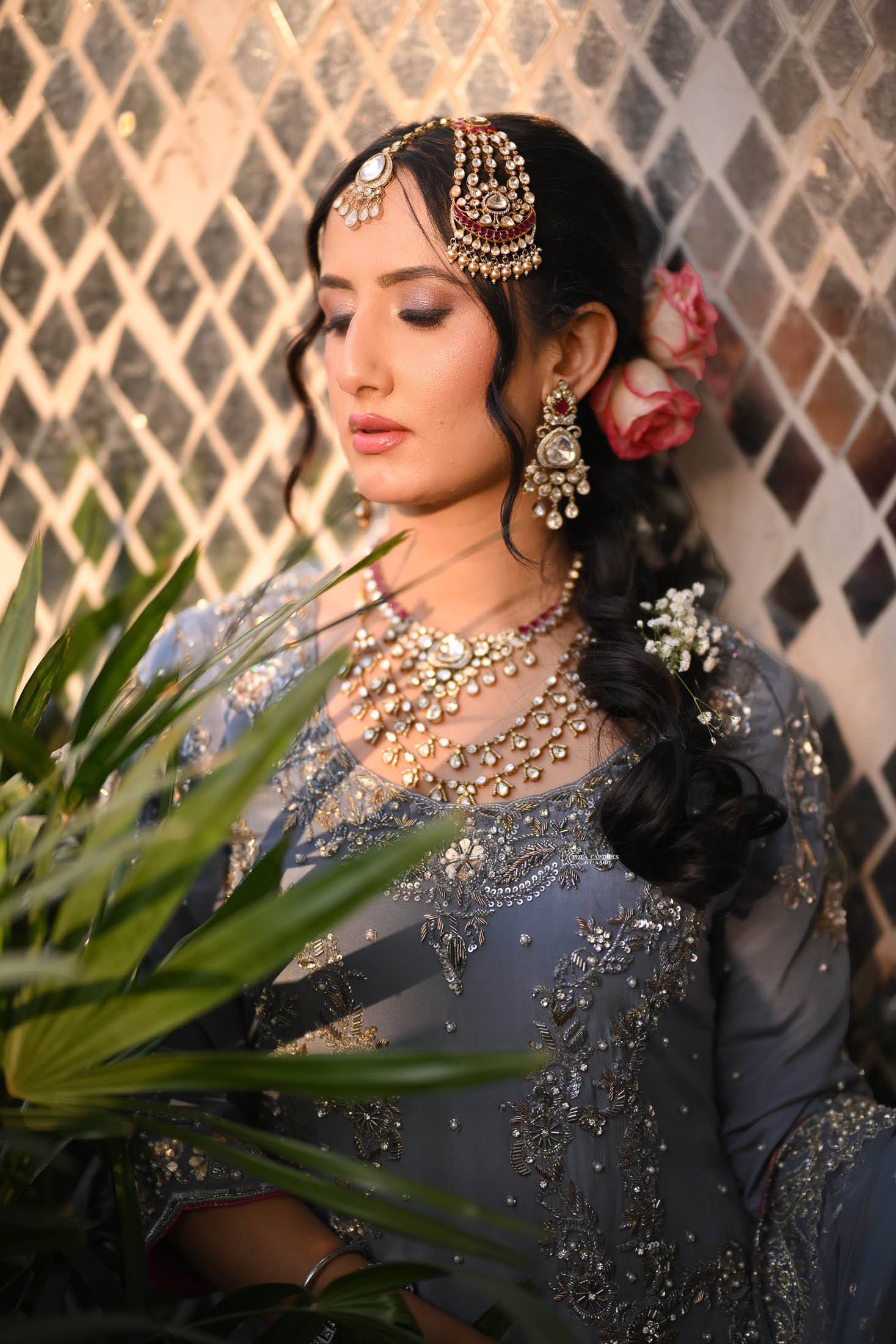 A woman dressed in traditional Indian attire with elaborate jewelry, including earrings, necklaces, and a headpiece, standing near a plant with a lattice background.