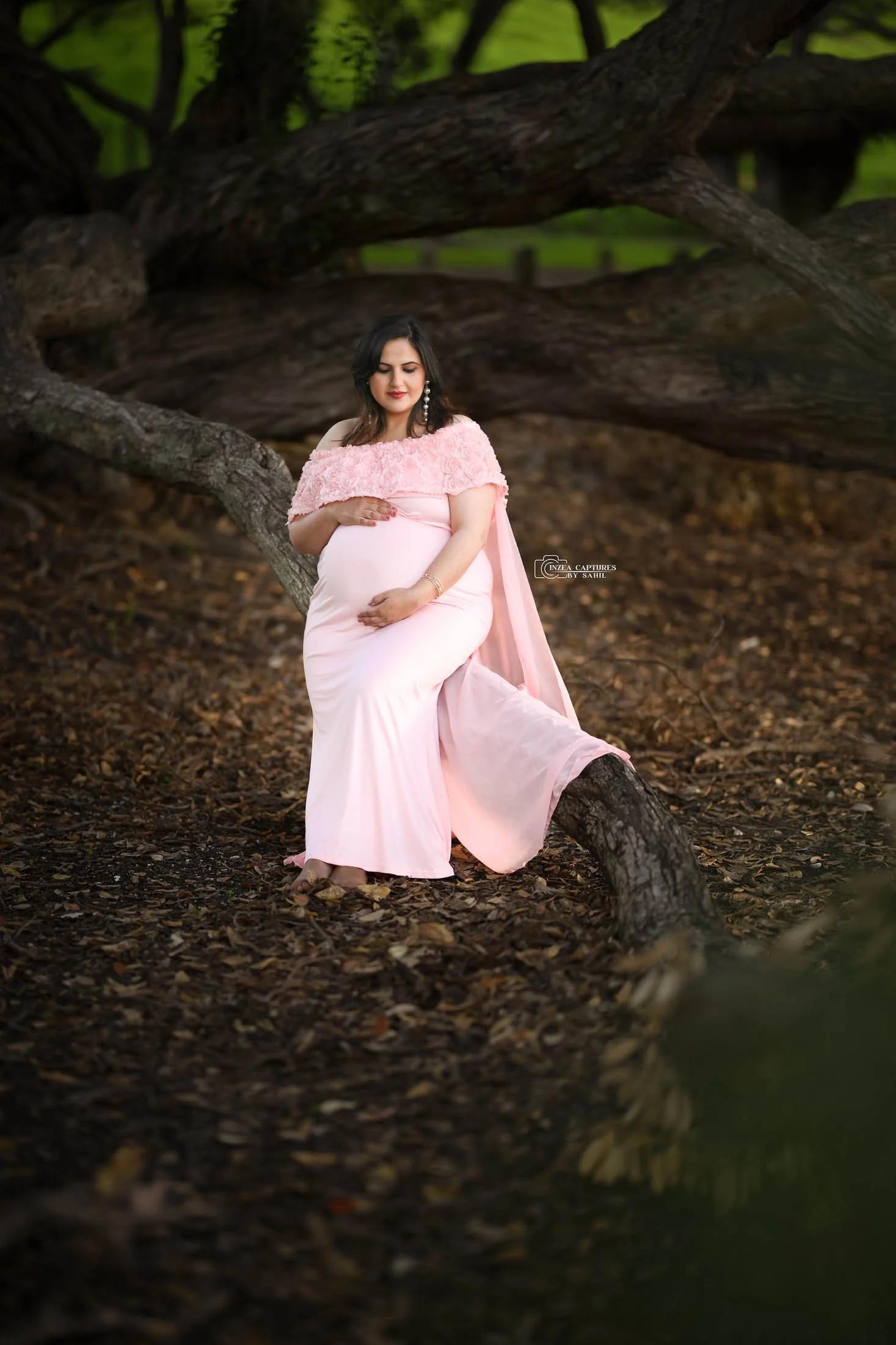 A pregnant woman in a pink dress sitting on a tree branch in a forest.