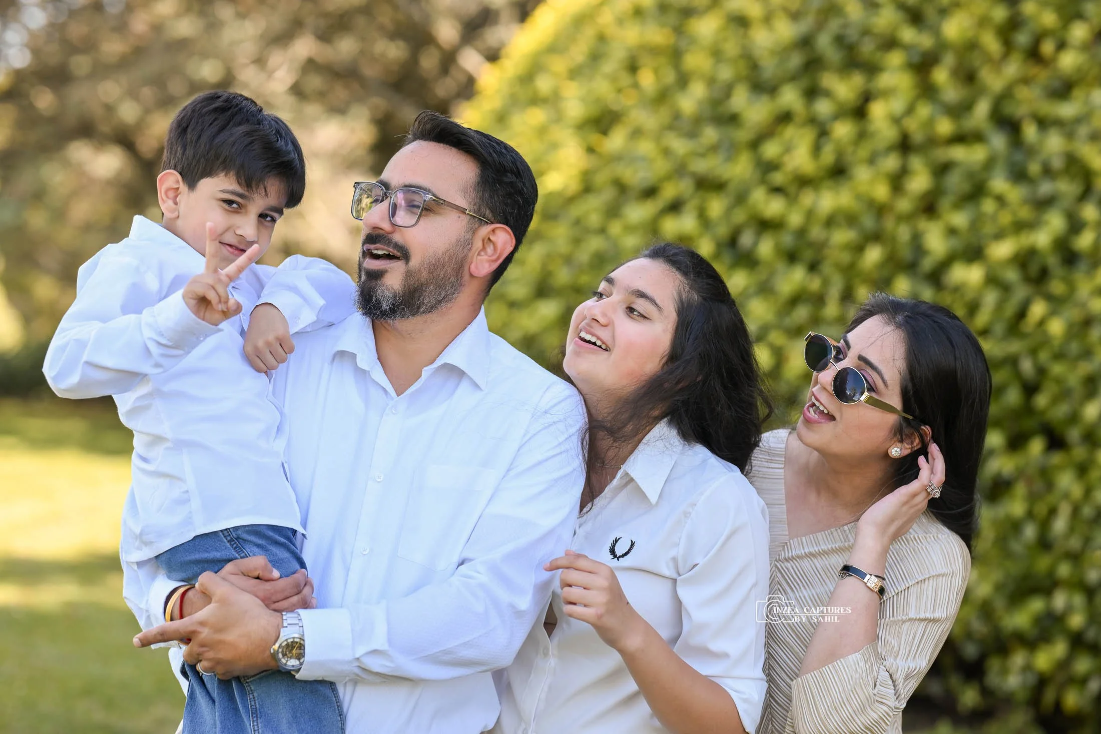 A family of four enjoying time outdoors in a park, with trees in the background. The father is holding a young boy, who is making a peace sign, while a woman and another girl watch and smile.