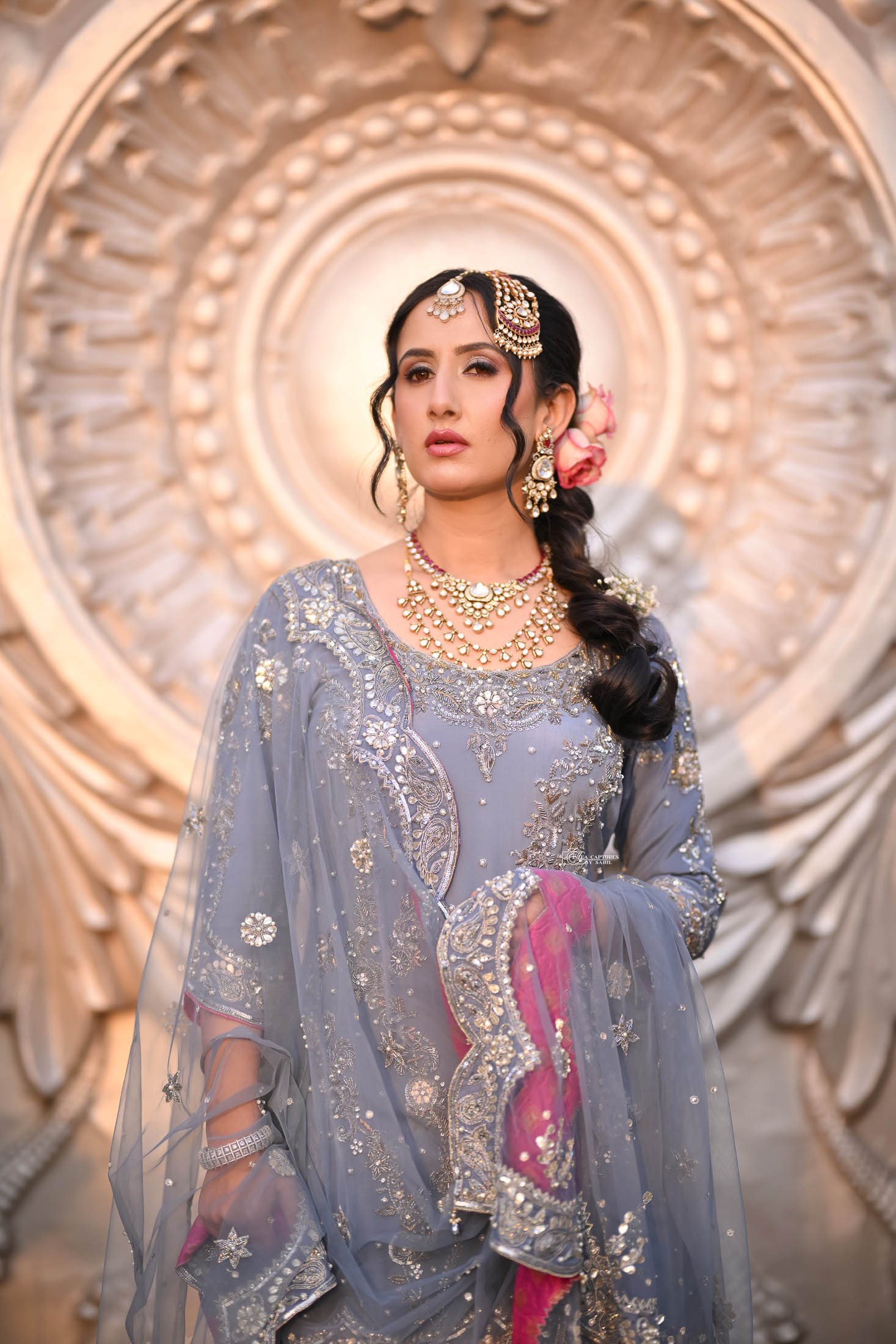 A woman dressed in an elegant traditional grey and pink embroidered outfit, adorned with gold jewelry, including necklaces, earrings, and a maang tikka, standing in front of an ornate background.