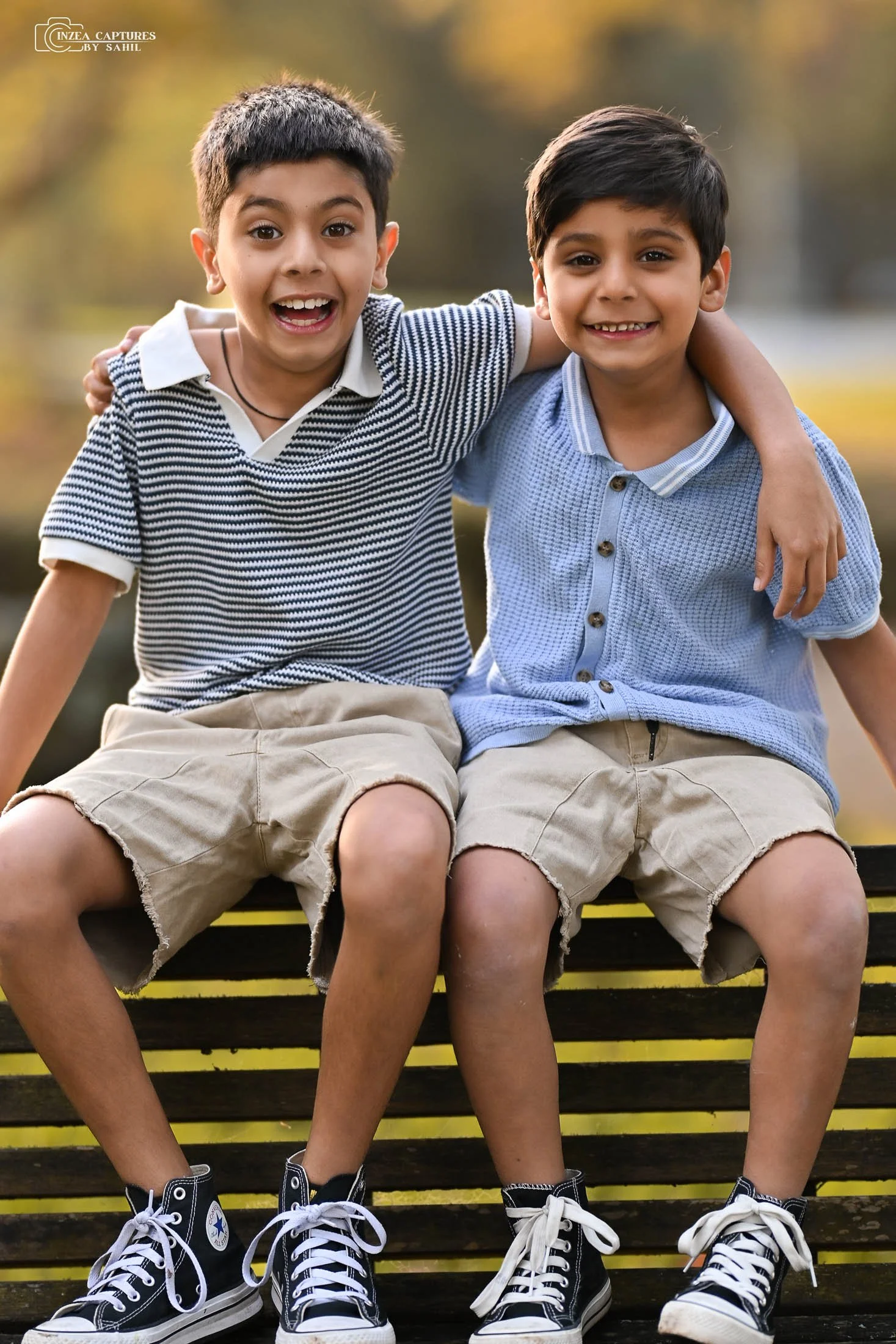 Two young boys sitting on a park bench outdoors, smiling, with arms around each other, wearing casual summer clothes and sneakers, with autumn trees in the background.