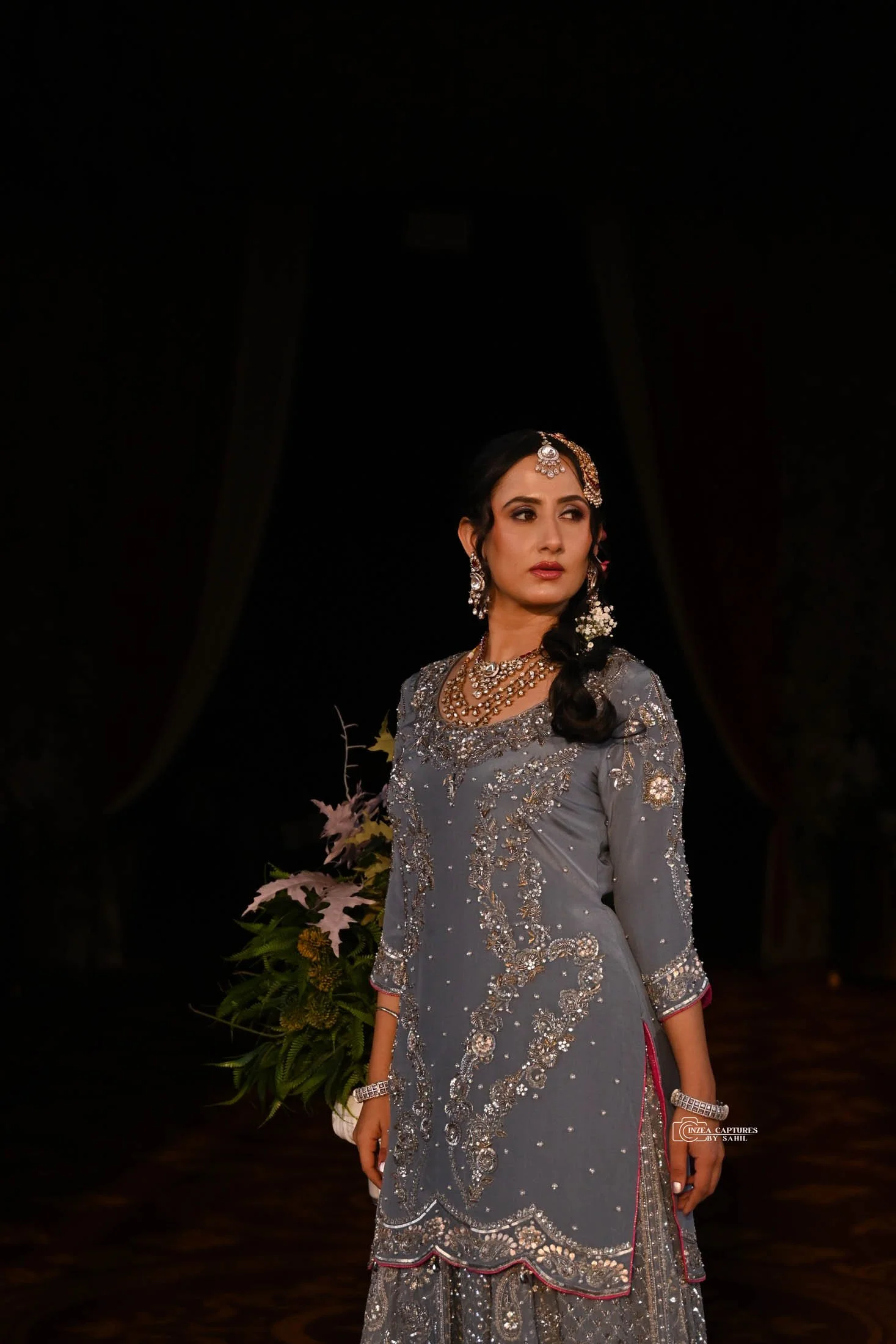 A woman dressed in a traditional ornate gray dress with silver embellishments, standing indoors at night with a dark background and a floral arrangement in the background.