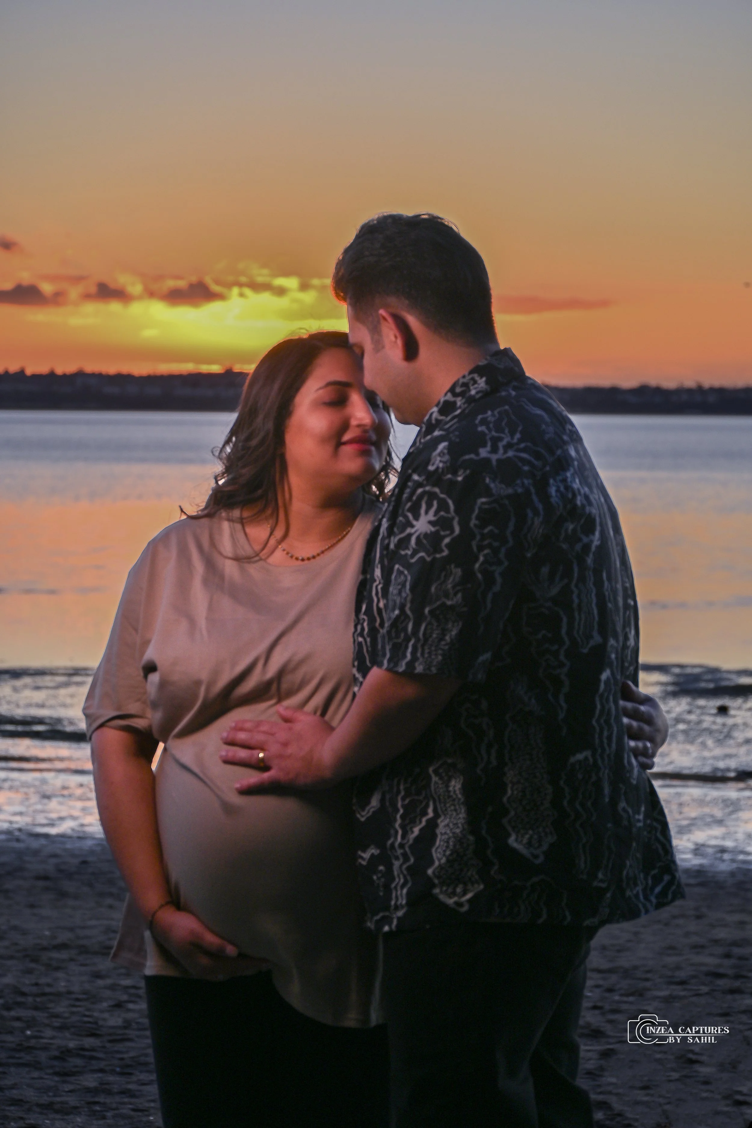 A couple stands close together on a beach at sunset, with the woman visibly pregnant, sharing an intimate moment by the water.
