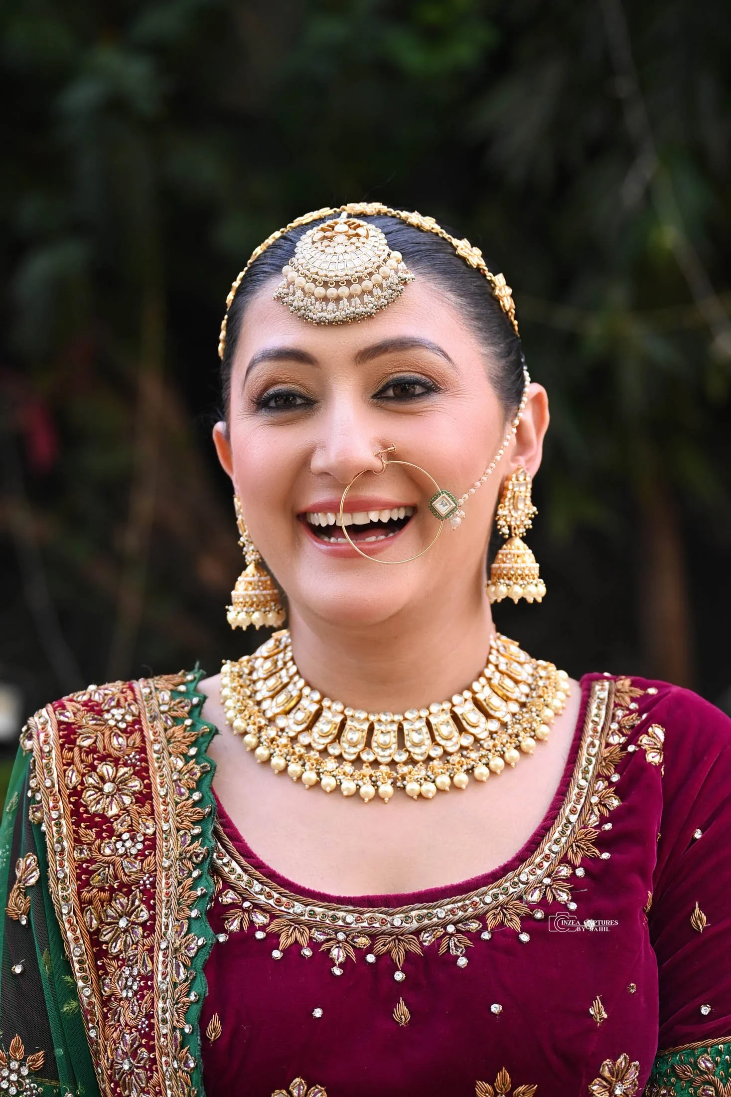 A woman dressed in traditional Indian attire, wearing ornate jewelry including a large necklace, earrings, a headpiece, and a nose ring with a chain. She is smiling and appears to be outdoors.