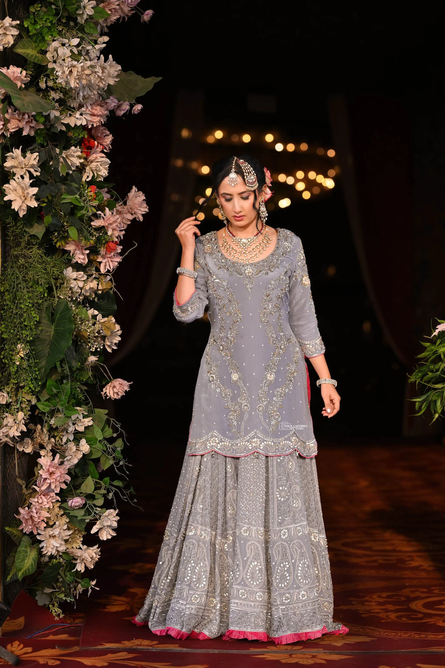 A woman in an ornate silver traditional dress decorated with embroidery and embellishments, standing on a dark stage with floral arrangements and warm lighting in the background.