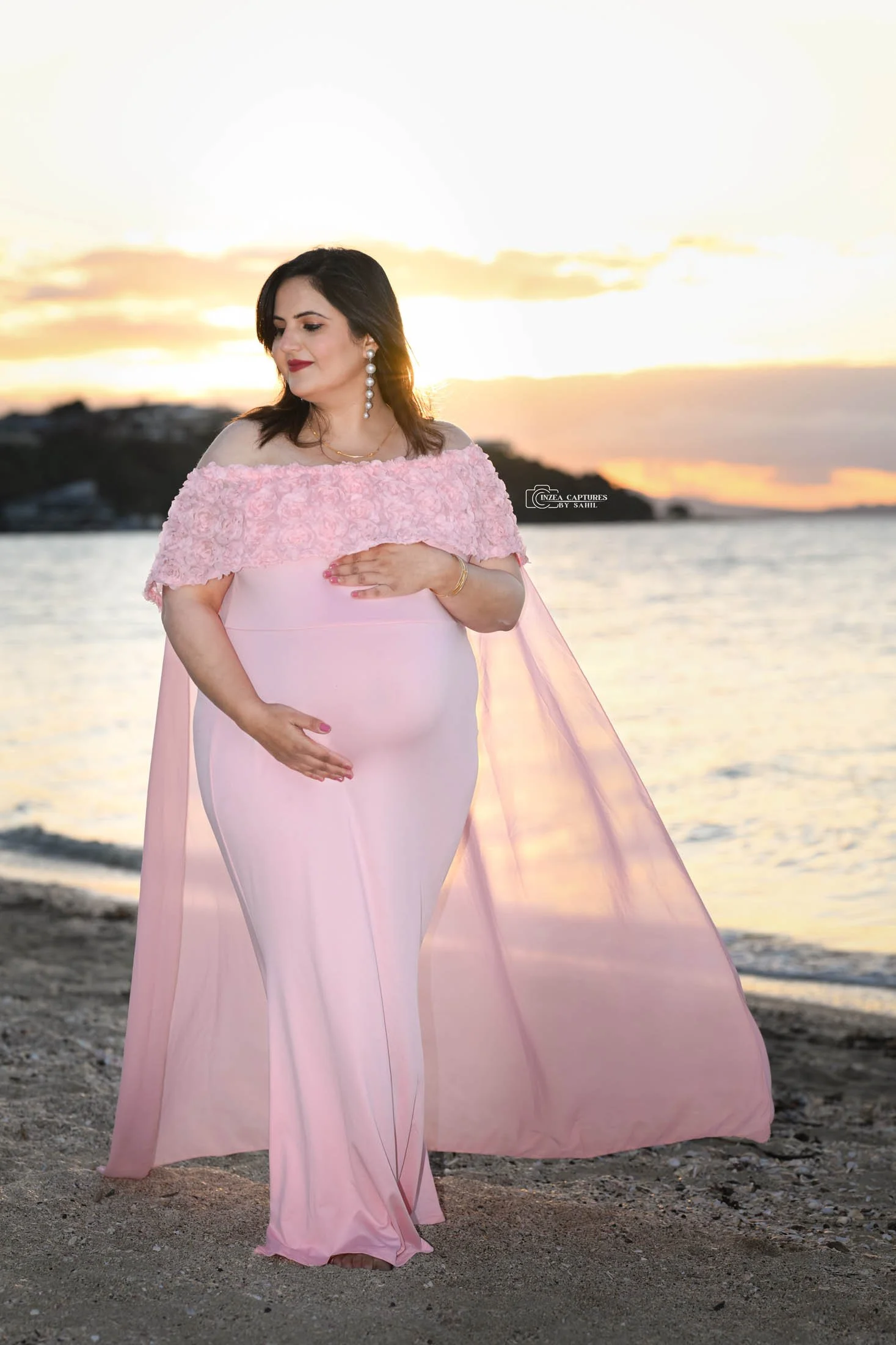 A pregnant woman in a pink gown with a floral off-shoulder overlay, standing on a beach during sunset, gently holding her belly with one hand and the other on her chest, with earrings and a bracelet.