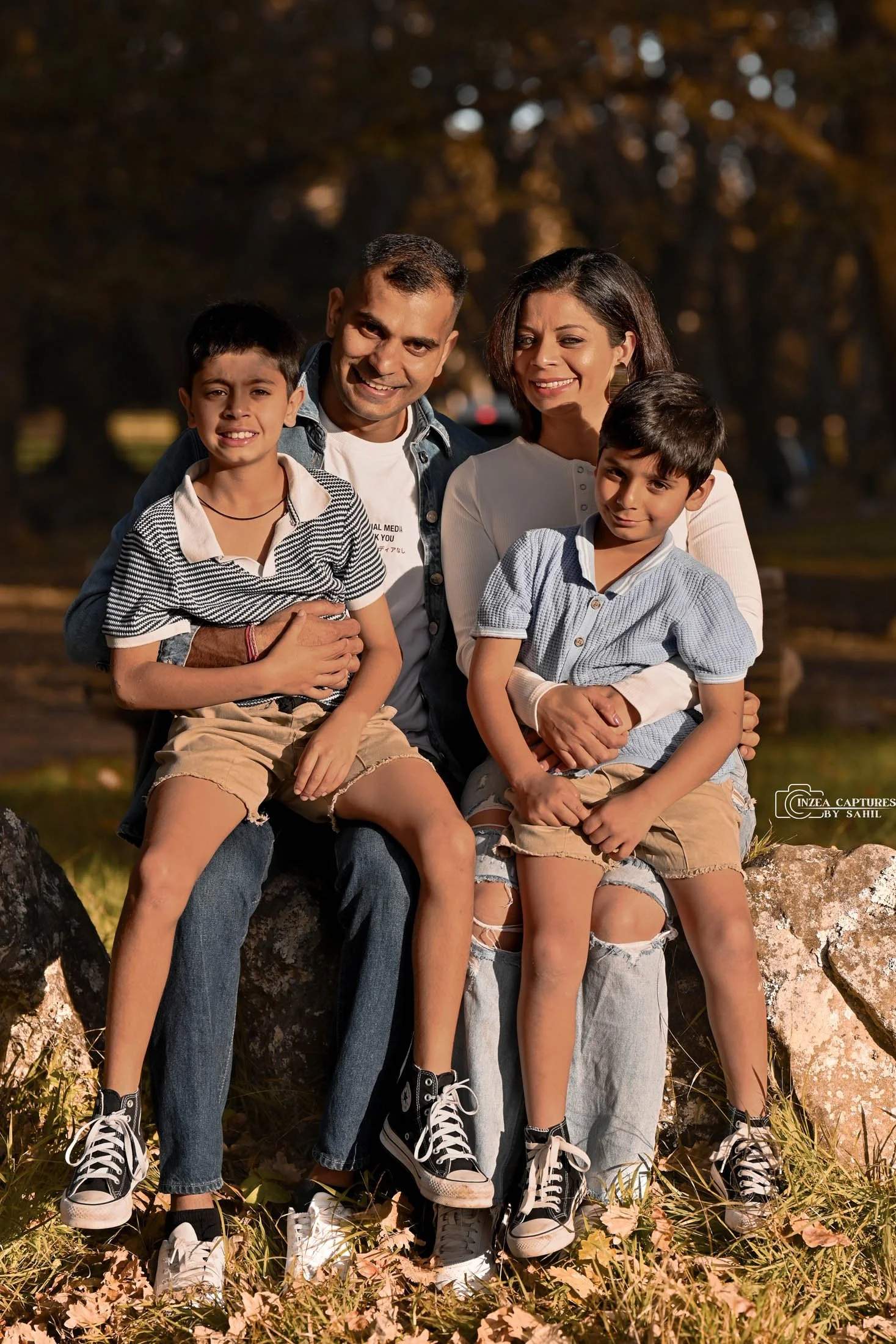 A happy family of four sitting on rocks outdoors in autumn, smiling at the camera. The father and mother sit behind their two sons, who are wearing casual clothing and sneakers.