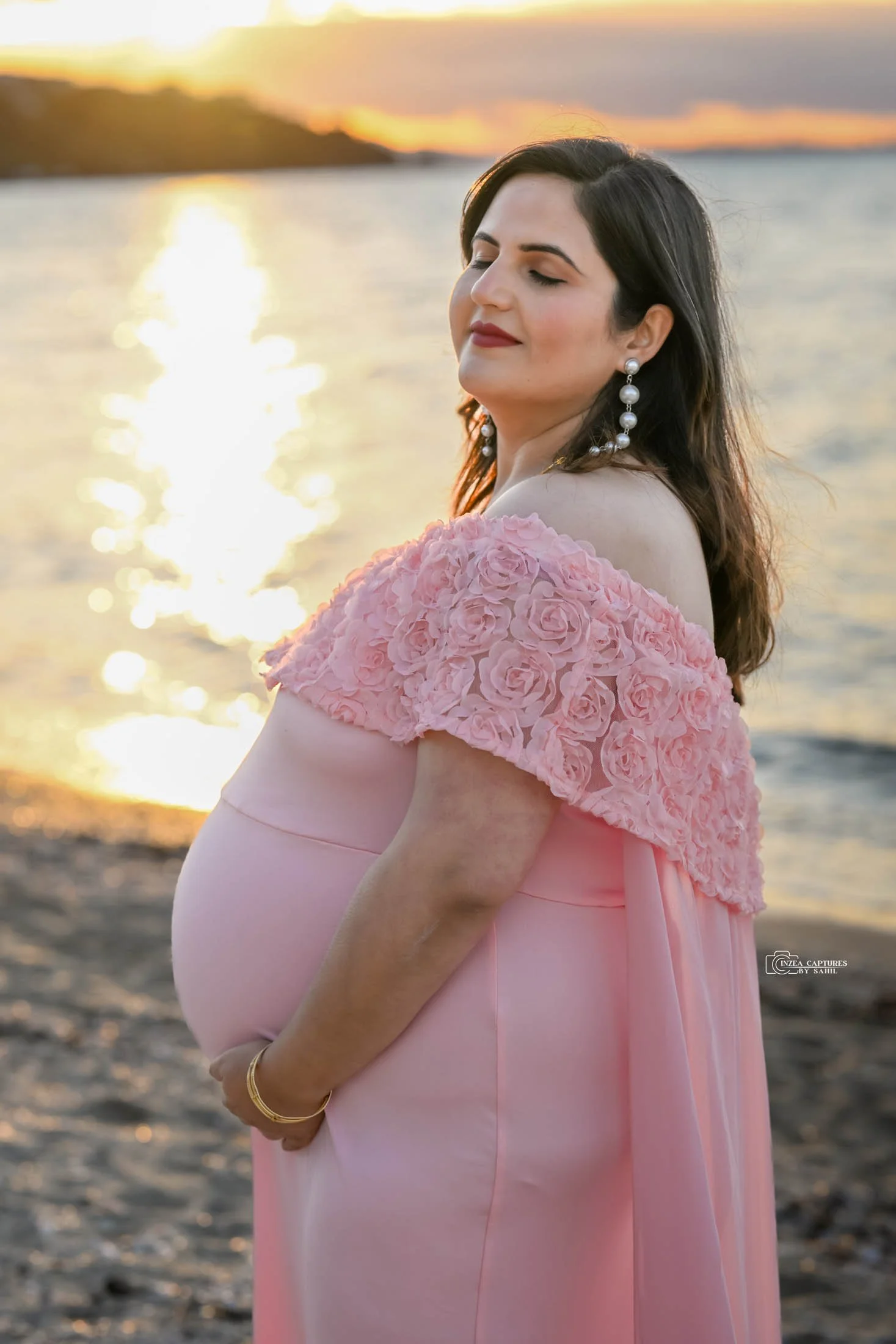 A woman in a pink dress with floral embellishments on the shoulders stands by the water during sunset, with closed eyes and a serene expression.