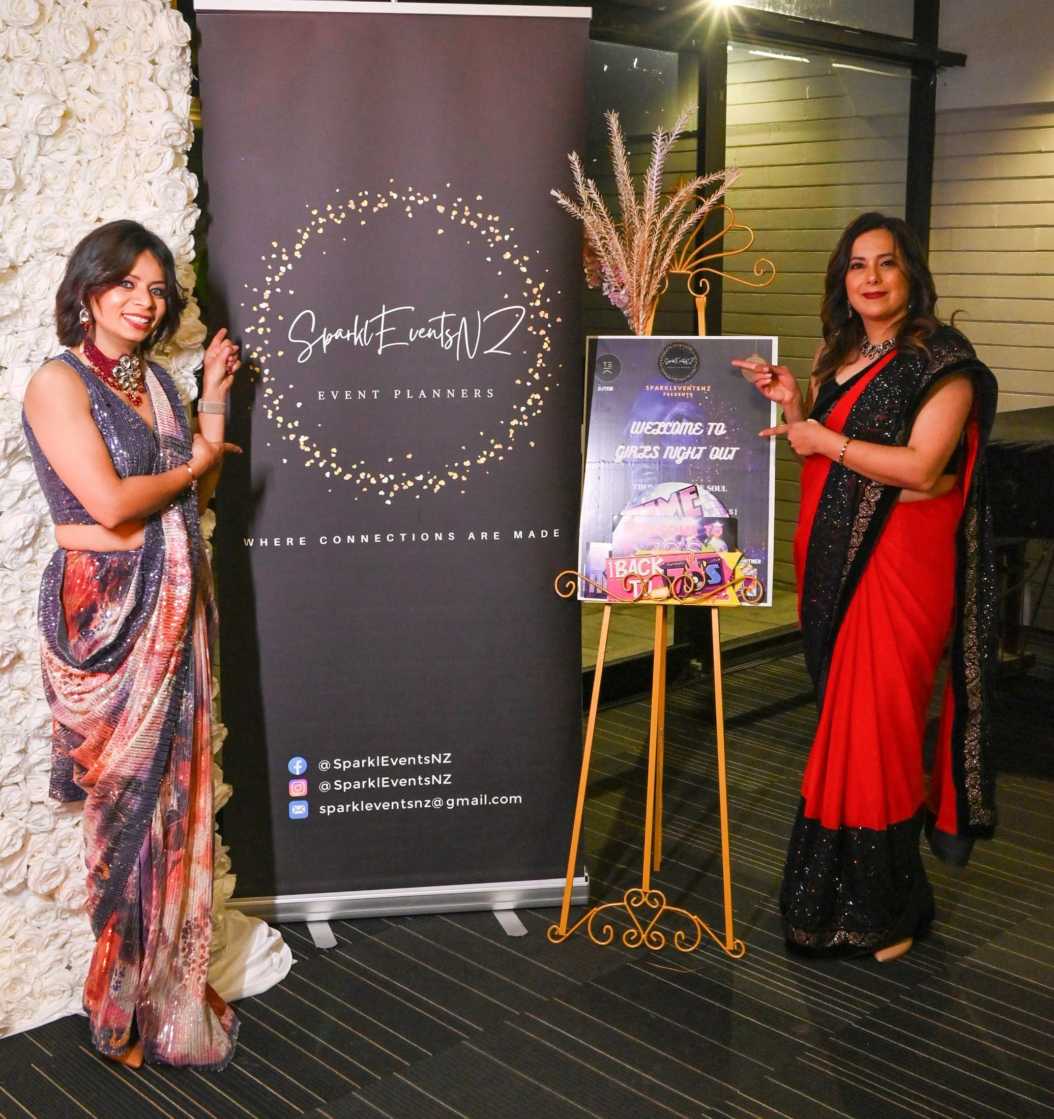 Two women dressed in colorful sarees at a formal event, standing next to a black banner for SparkleEventsNZ. One woman on the left points to the banner, while the other on the right holds a sign with event details. The banner displays the logo and so