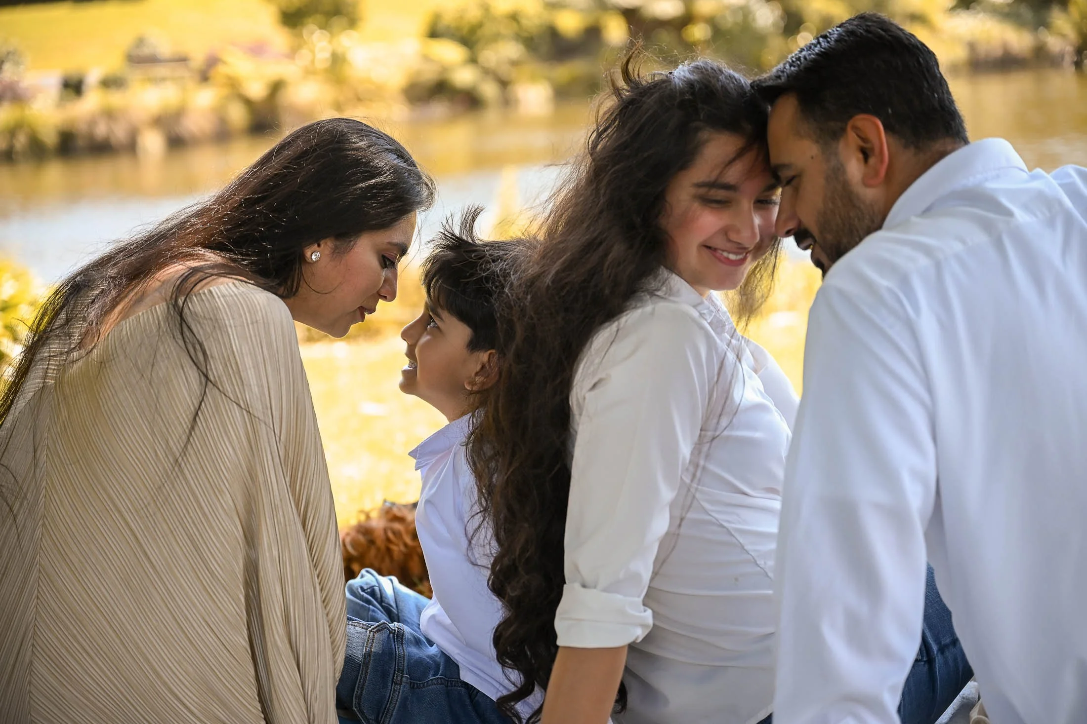 A family of four outdoors by a lake, sitting closely with foreheads touching, smiling and enjoying each other's company.