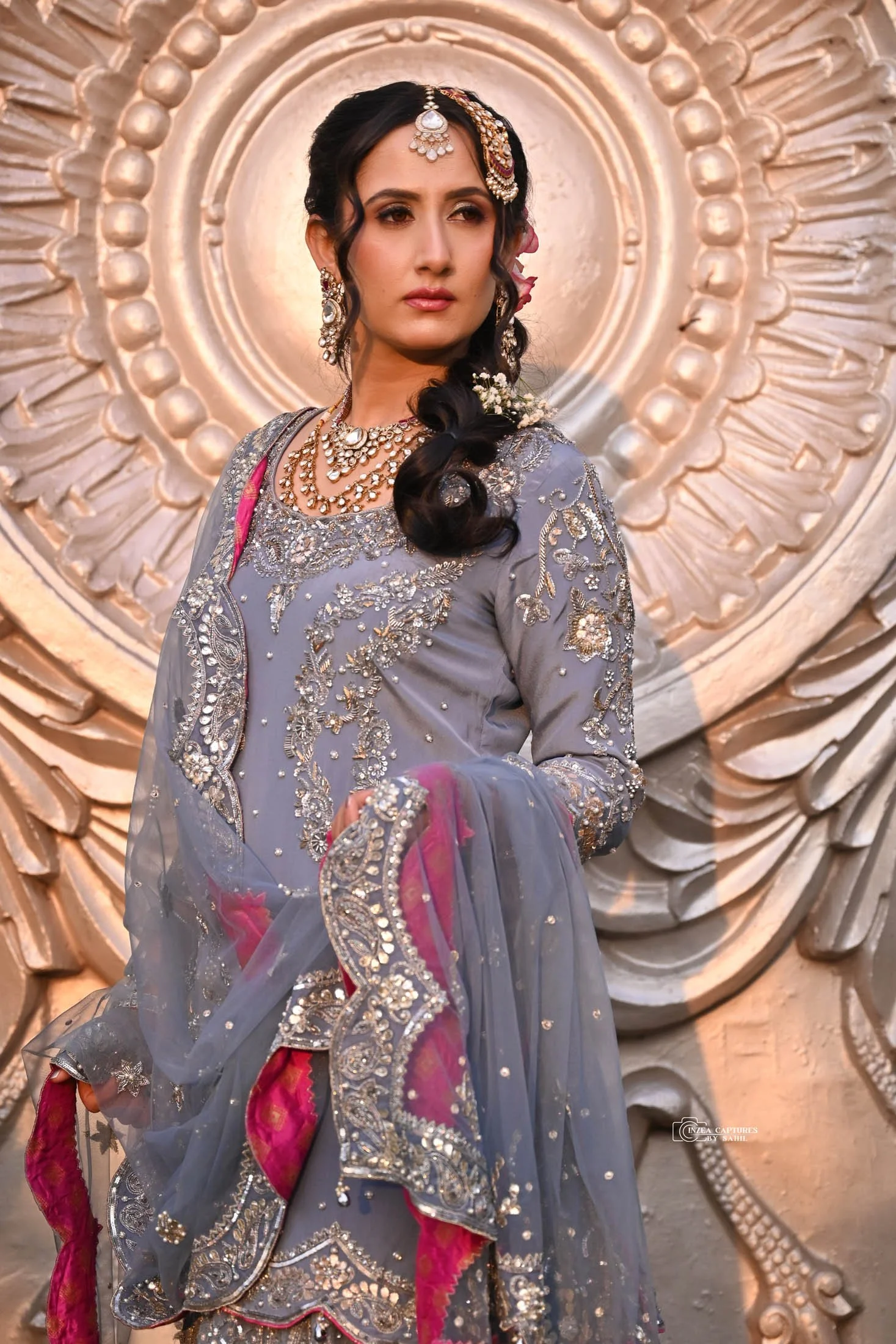 A woman in traditional Indian attire, wearing a gray embroidered dress with pink accents, and adorned with silver jewelry including earrings, necklaces, and a maang tikka, poses in front of a decorative backdrop with intricate circular patterns.