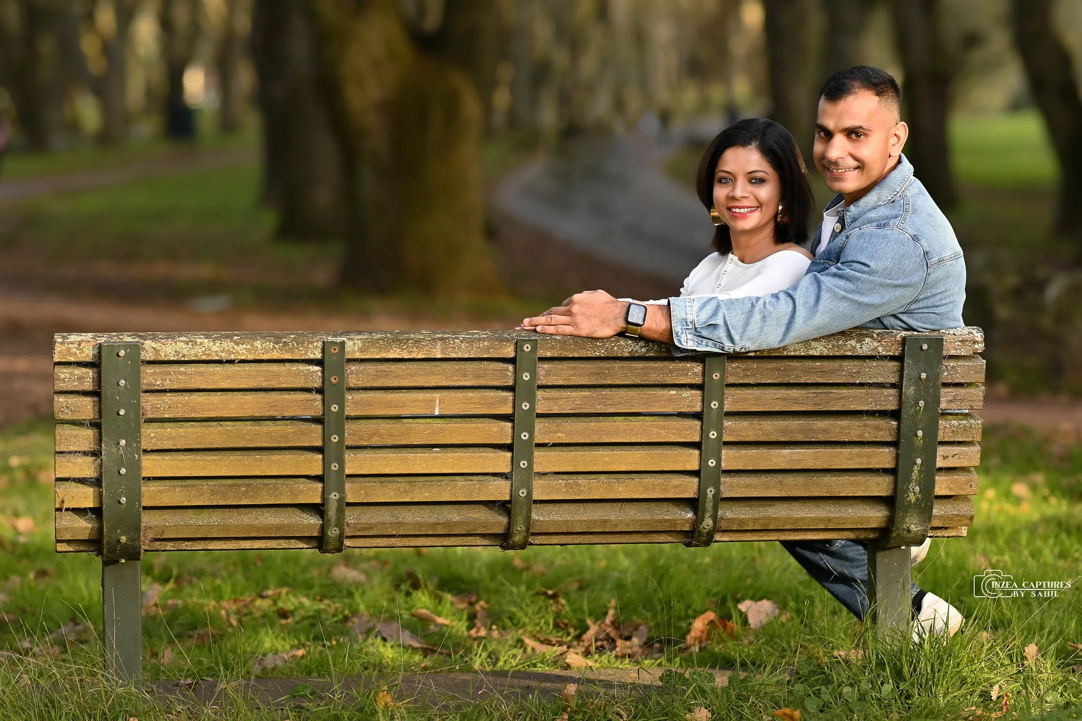 A smiling couple sitting on a park bench, with trees and a walking path in the background.