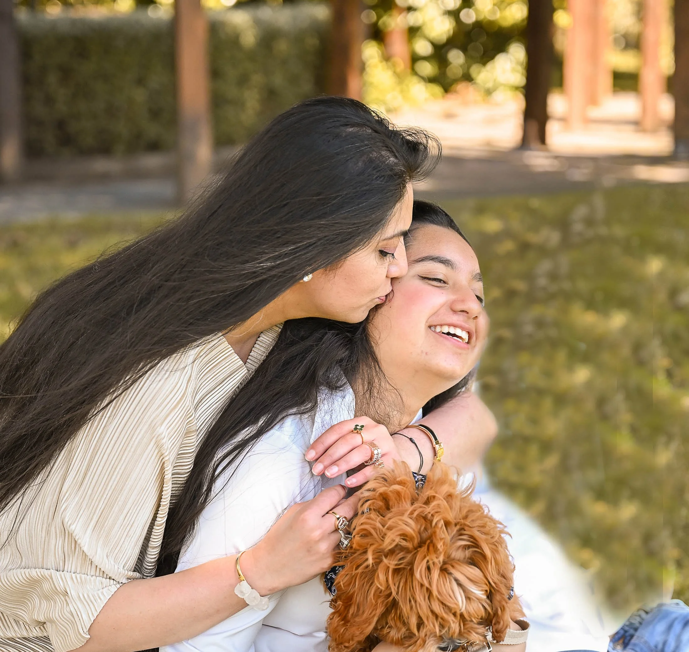 Two women sharing a loving moment outdoors near a body of water, with one woman kissing the other's head and holding a small brown dog.
