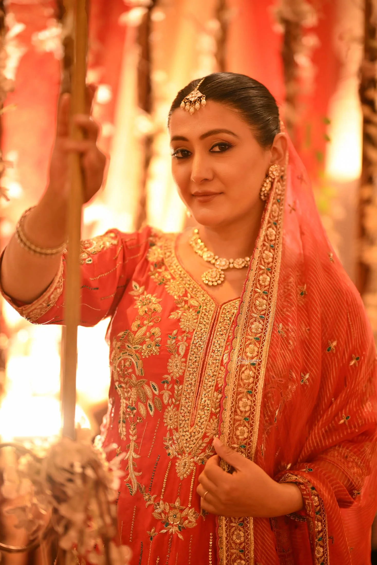 A woman dressed in traditional Indian attire, wearing an orange saree with gold embroidery, jewelry, and a veil, standing in a decorated setting with warm lighting.