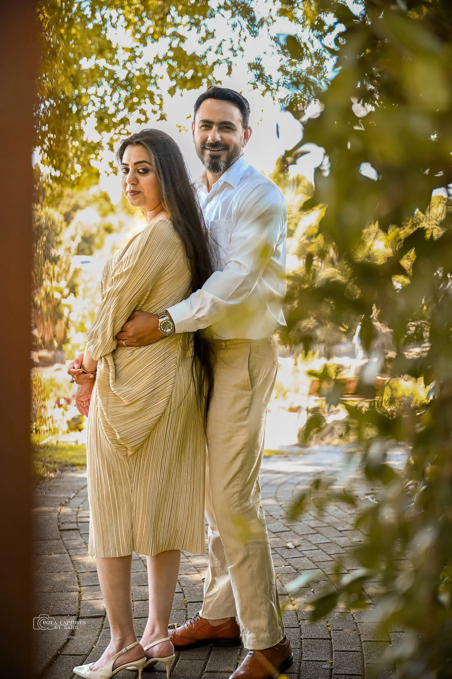 A couple standing outdoors on a brick path surrounded by greenery, with sunlight filtering through the leaves, the woman wears a beige pleated dress and heels, and the man wears a white shirt and beige pants, both smiling softly.