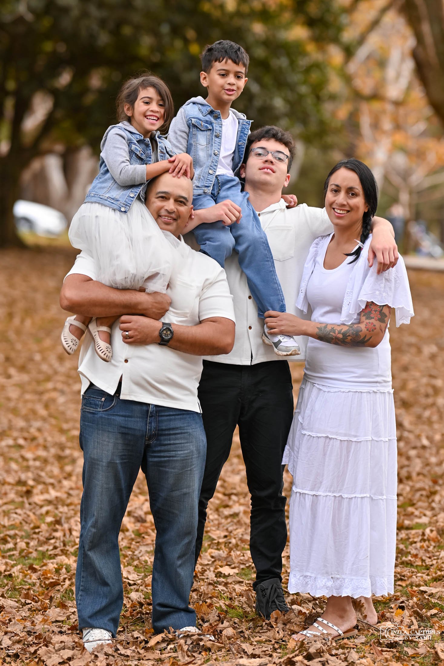 Family of five outdoors on a fall day with fallen leaves, smiling, with two children sitting on parents' shoulders.