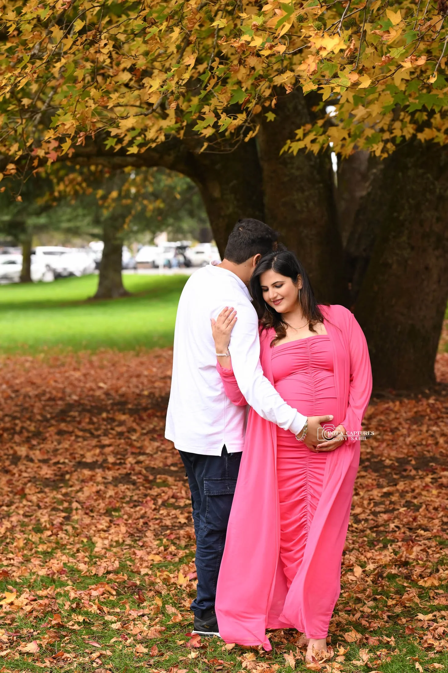 A pregnant woman in a pink dress and a man in a white shirt stand close together under a large tree with palm leaves, with fallen leaves on the ground surrounding them.