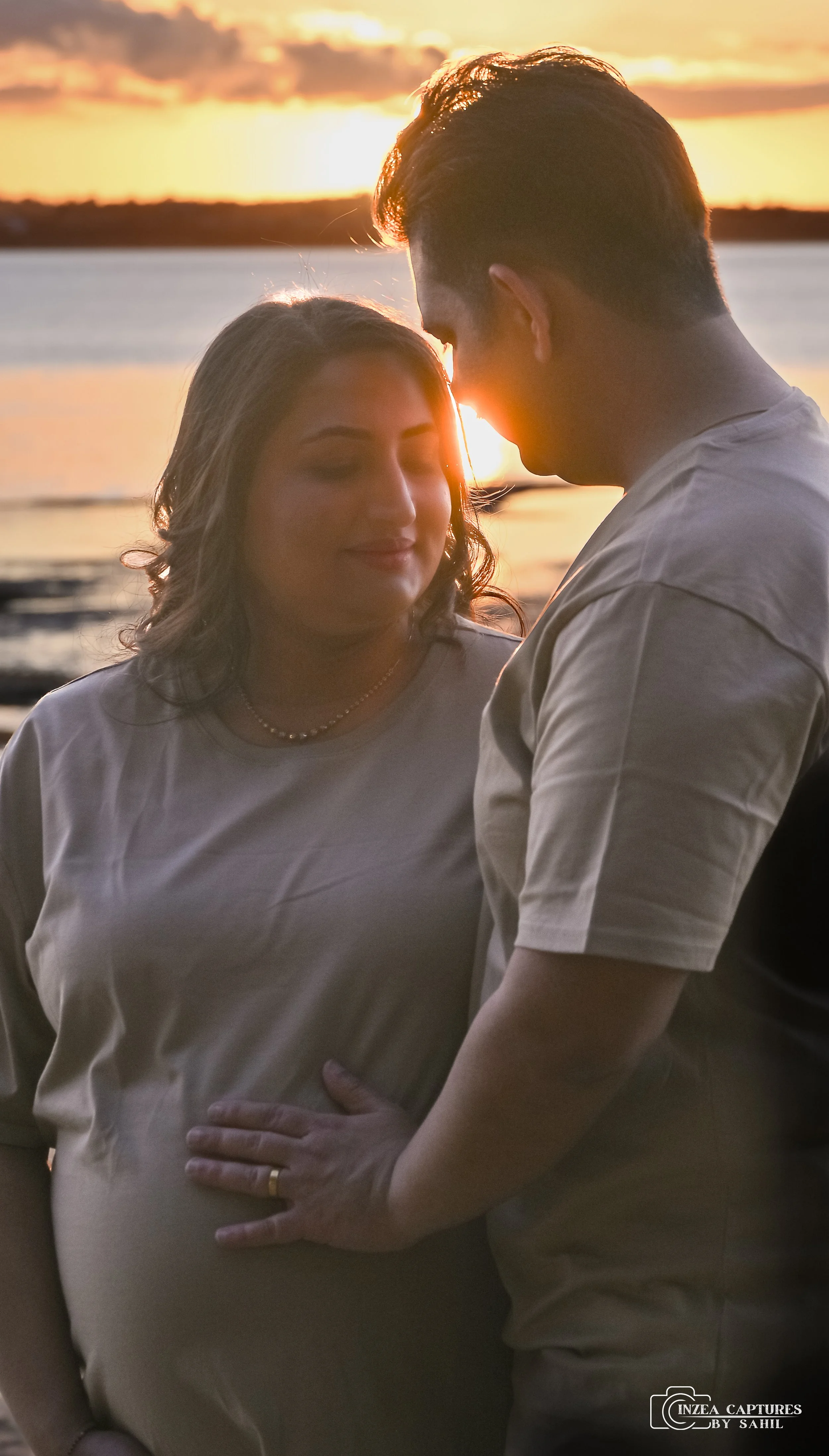 A couple standing close together during sunset by a body of water, with the man gently resting his hand on the woman's pregnant belly, both smiles softly.