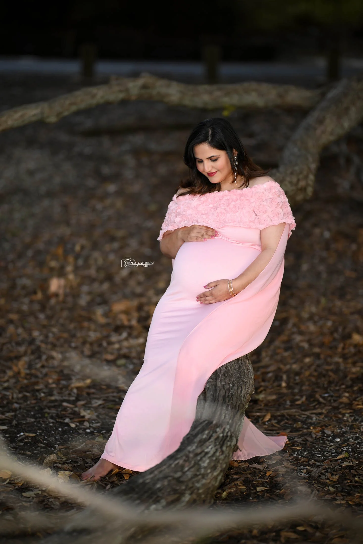 Pregnant woman in pink dress sitting on a fallen tree in a wooded area, looking down and touching her belly.