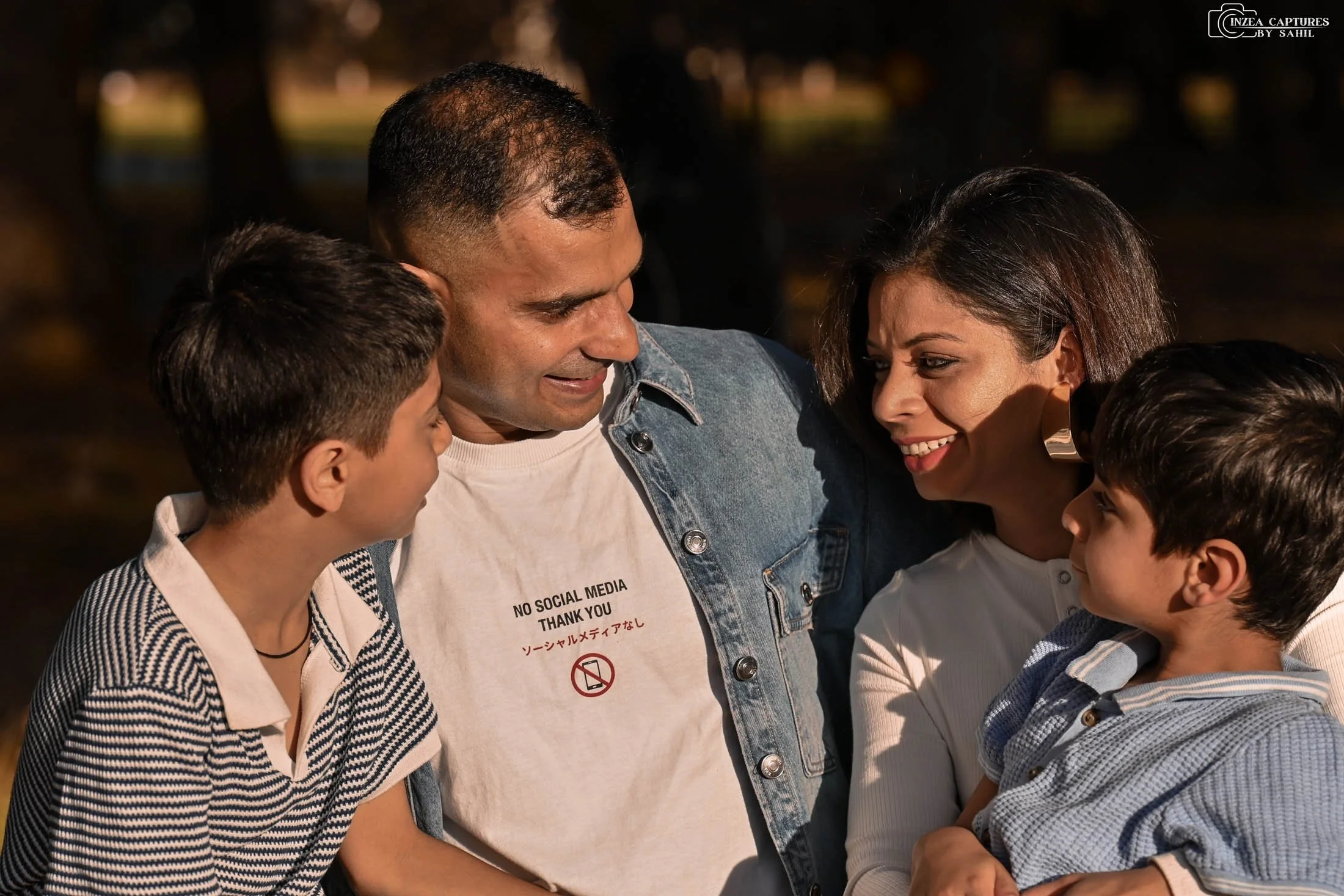 A family of five smiling and looking at each other outdoors at night, with trees in the background, lit by warm light.