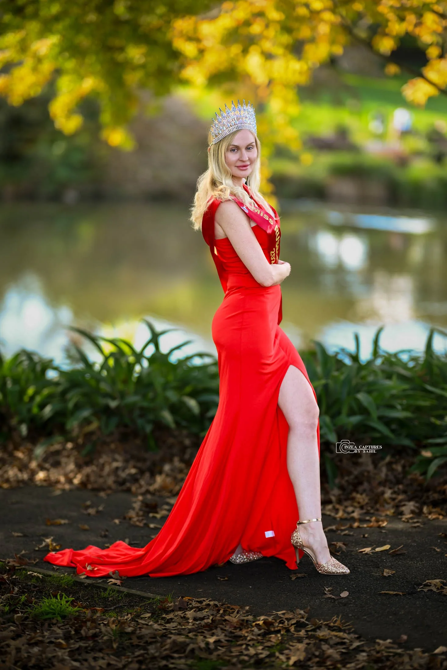 A woman in a red gown with a high slit, wearing a crown and sash, standing outdoors near a lake with autumn foliage in the background.