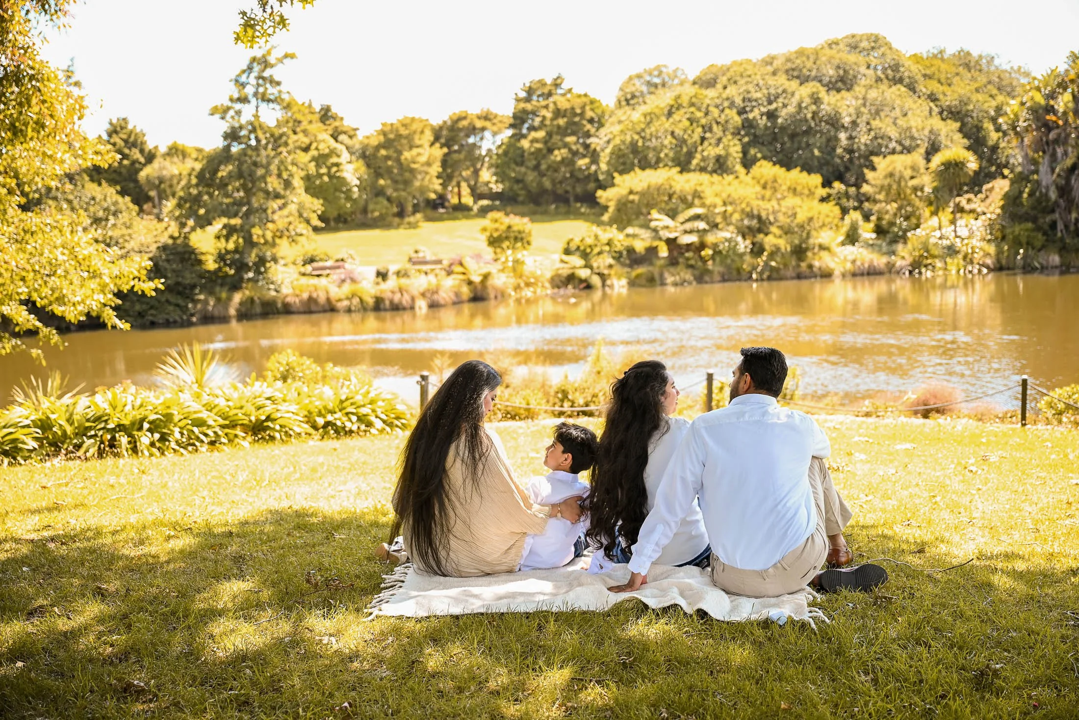 A family of four sitting on a blanket by a lake on a sunny day. The scene includes trees, grass, and a tranquil water body. The family appears to be enjoying a relaxing outdoor gathering.
